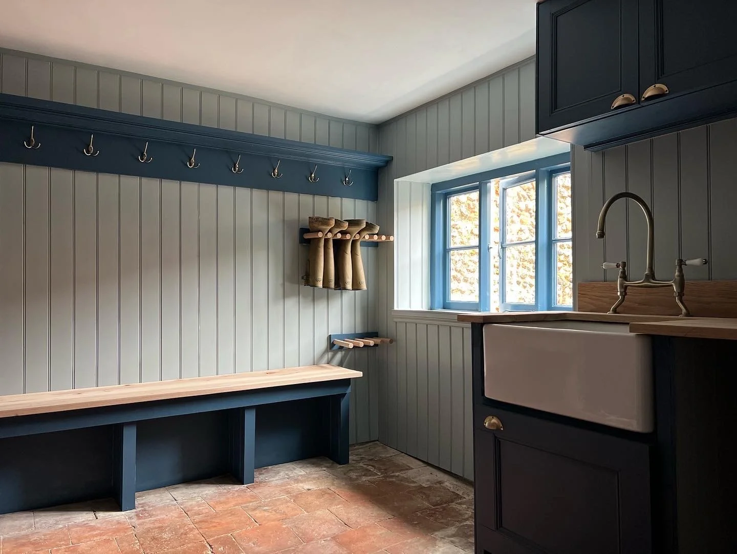 Interior of a rustic boot room with beadboard walls, an oak bench, coat hooks, a window with blue trim, and a farmhouse sink with brass fixtures.