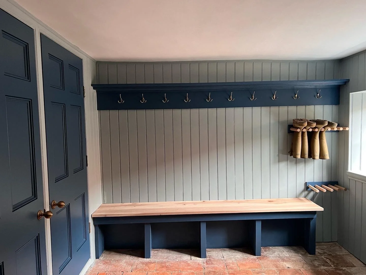 A boot room with a navy blue coat rack with hooks, an oak bench, and shoe storage with cubbie holes underneath. There are wooden pegs on the right side for hanging boots, and the room has light blue beaded board walls and a window with white trim.