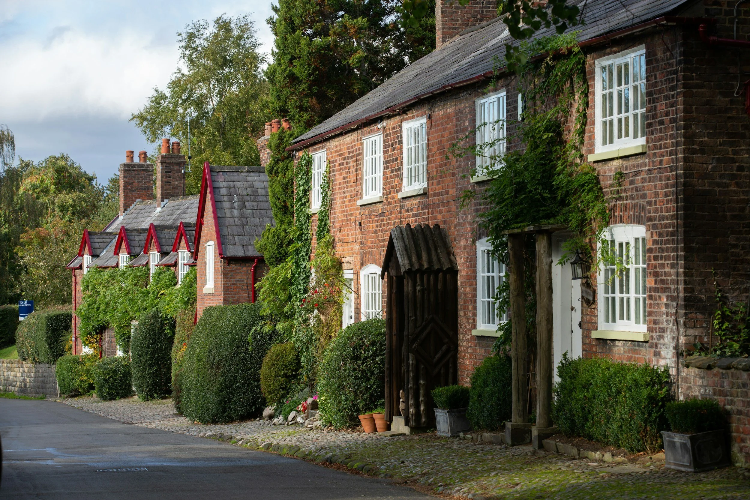 Row of brick houses with chimney stacks, white-framed windows, and greenery in front yards along a cobblestone sidewalk, under a partly cloudy sky.