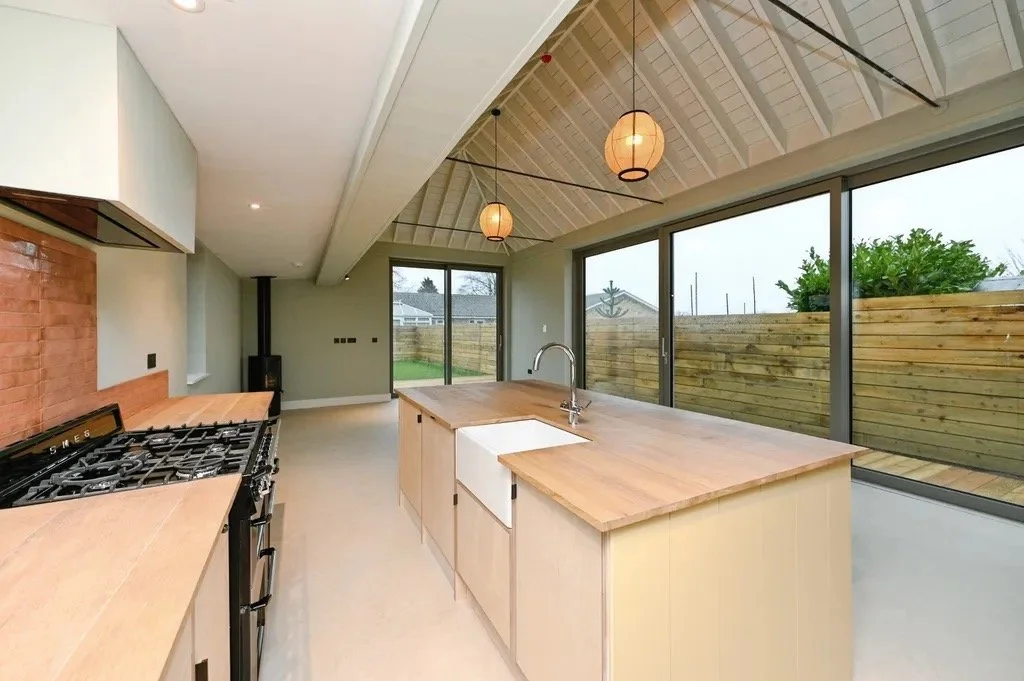 Modern kitchen with an island, wooden countertops, large glass sliding doors, and pendant lights hanging from a vaulted ceiling. There is an outdoor space visible through the doors.