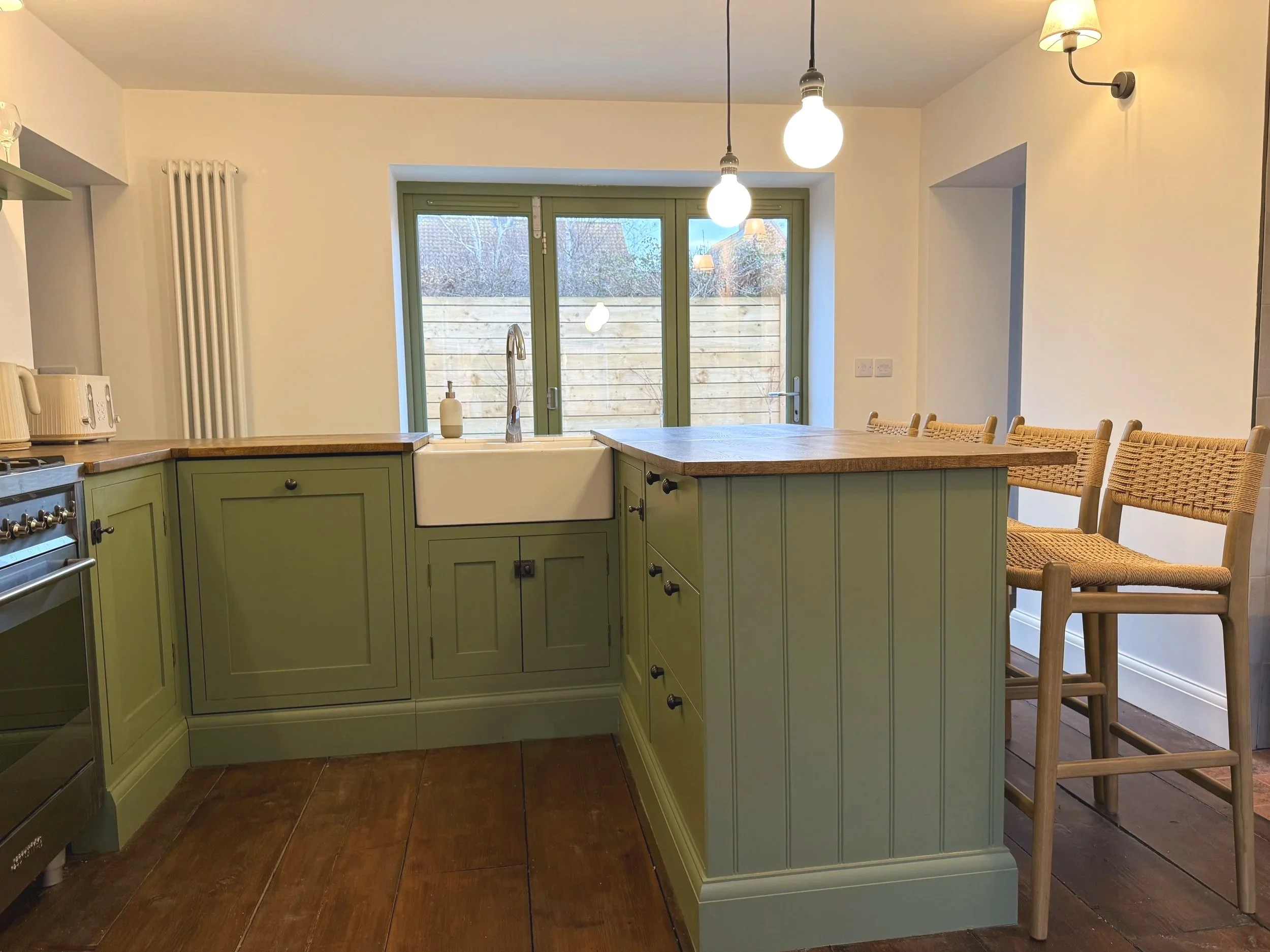 A handmade kitchen with green cabinets, a white farmhouse sink, and a wooden island with bar stools. There are two hanging porcelain pendant lights and an additional wall-mounted light, with large glass doors leading outside.