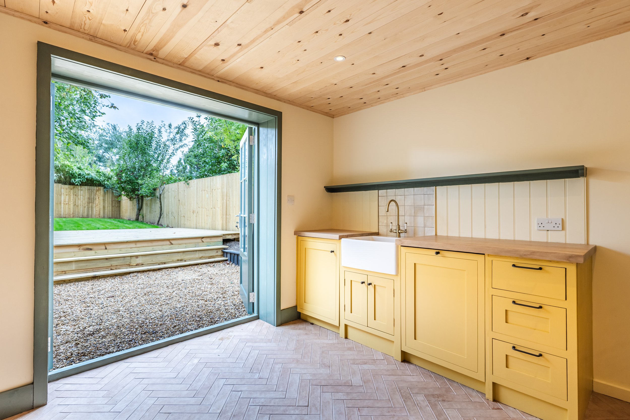 Interior view of a kitchen area with hand painted yellow cabinets, a double sink, and a window/door opening to a backyard with wooden steps, green grass, and trees.