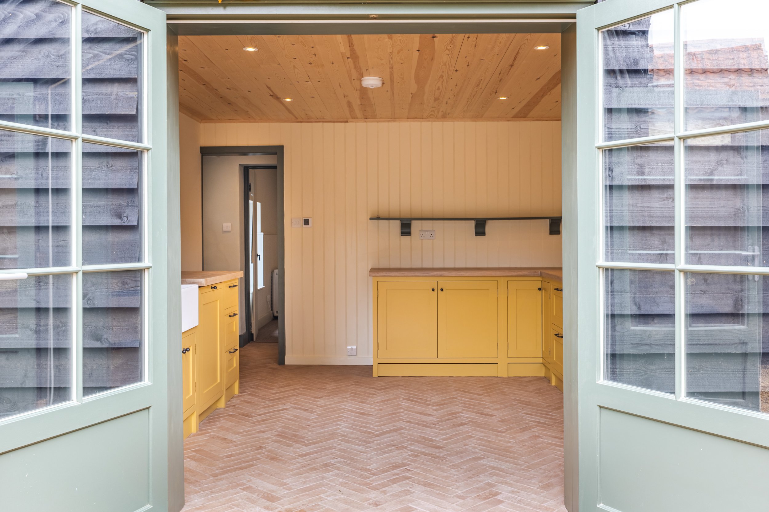 View of a kitchen with yellow cabinets and a wooden countertop, seen through open light green double doors with glass panes, wooden ceiling, beige wall, and herringbone terracotta tile flooring.