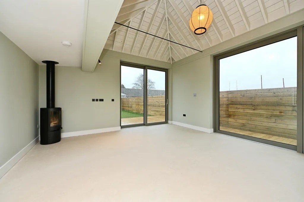 Empty living room with large glass sliding door and window, light-colored walls and flooring, black wood stove, and a pendant light hanging from a vaulted ceiling with exposed beams.