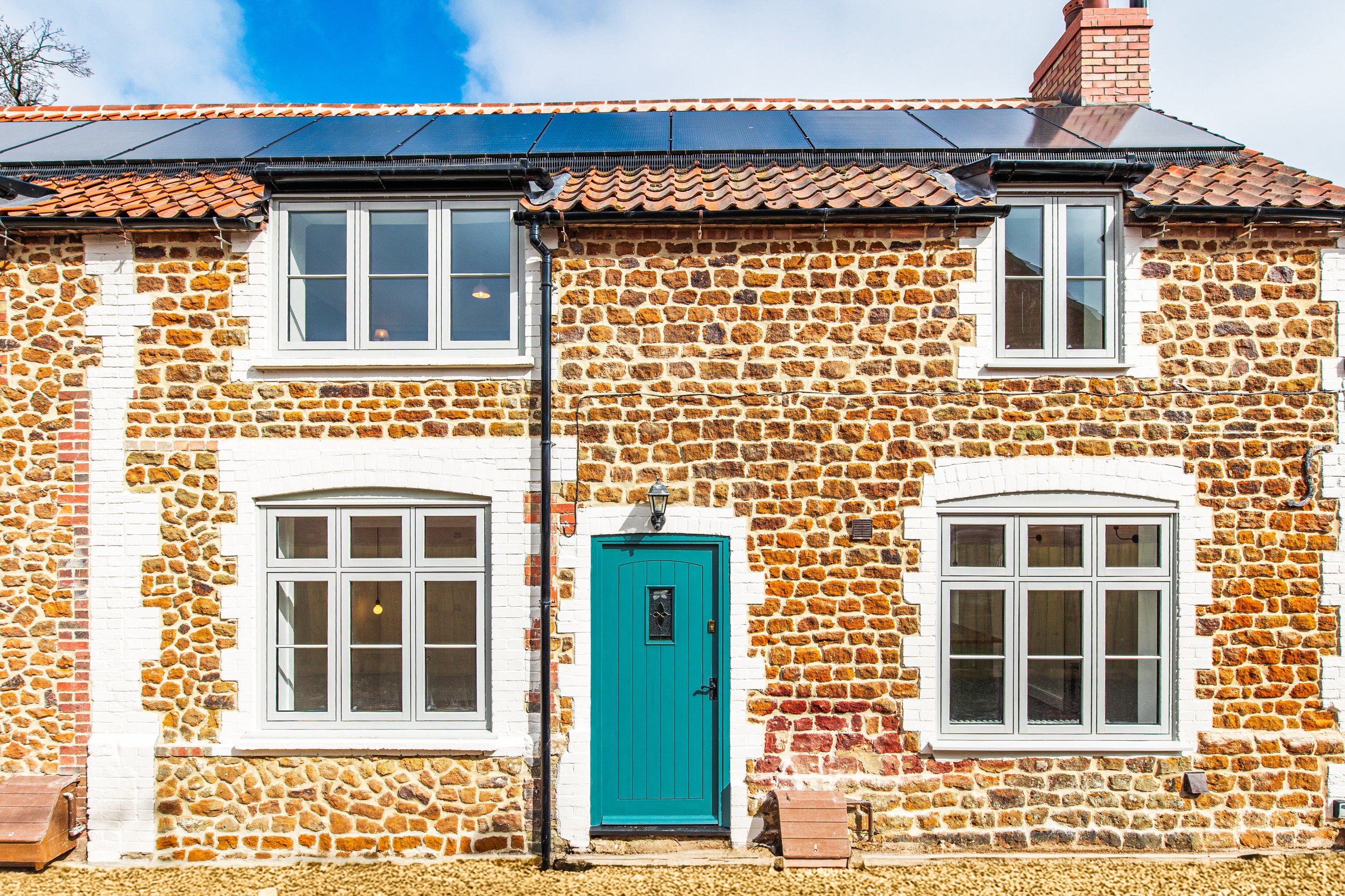 Front view of a two-story carrstone cottage with four Timberlook windows, a Paper & Paints front door, and solar panels on the roof.