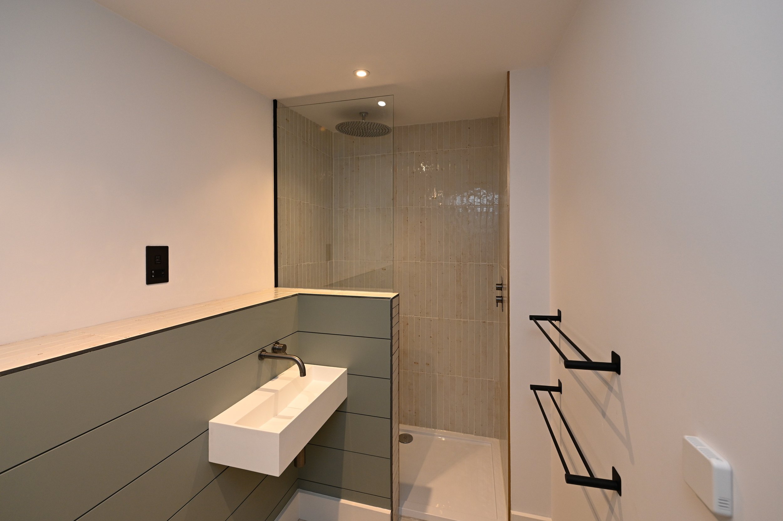 Modern bathroom with a walk-in shower, a wall-mounted sink with a black faucet, black towel bars, and a white wall with a black electrical outlet.