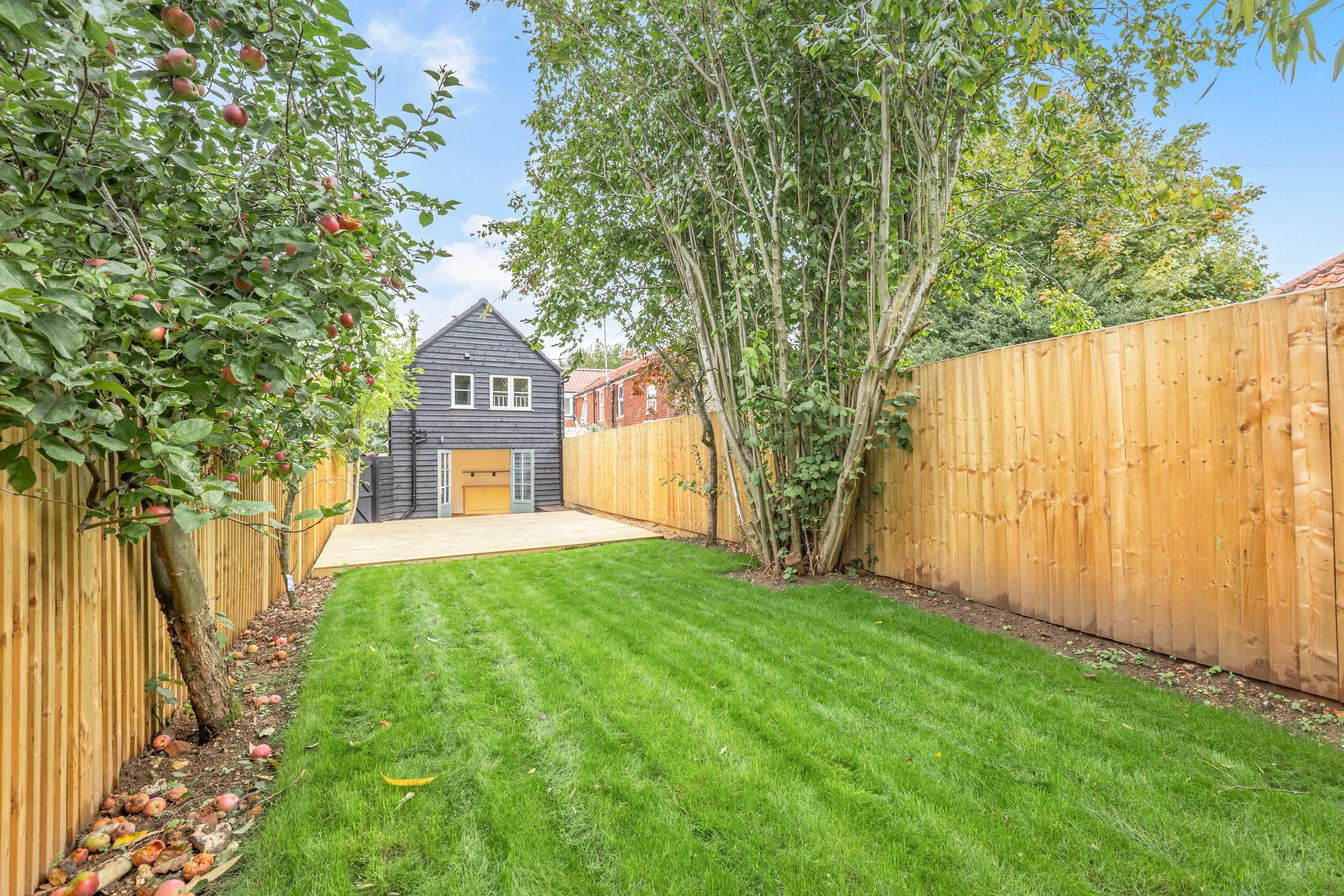 A backyard with a green lawn, trees, and a wooden fence, with a view of the timber-clad rear wall
