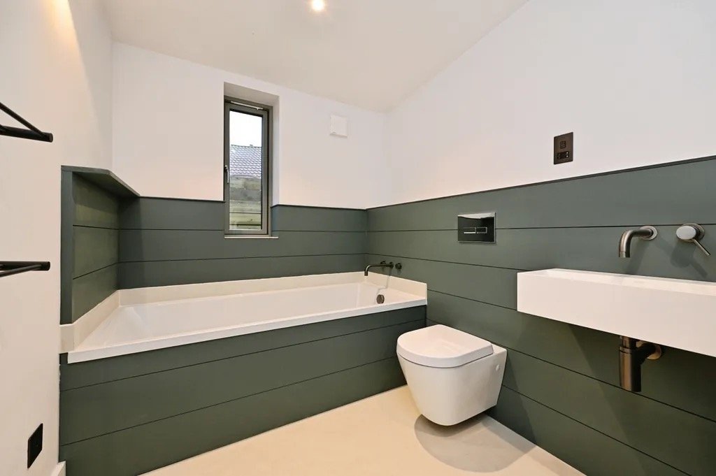 Modern bathroom with a bathtub, a toilet, and a minimalist sink. The walls have horizontal green and white paneling, a small window, and a black wall-mounted flush plate.