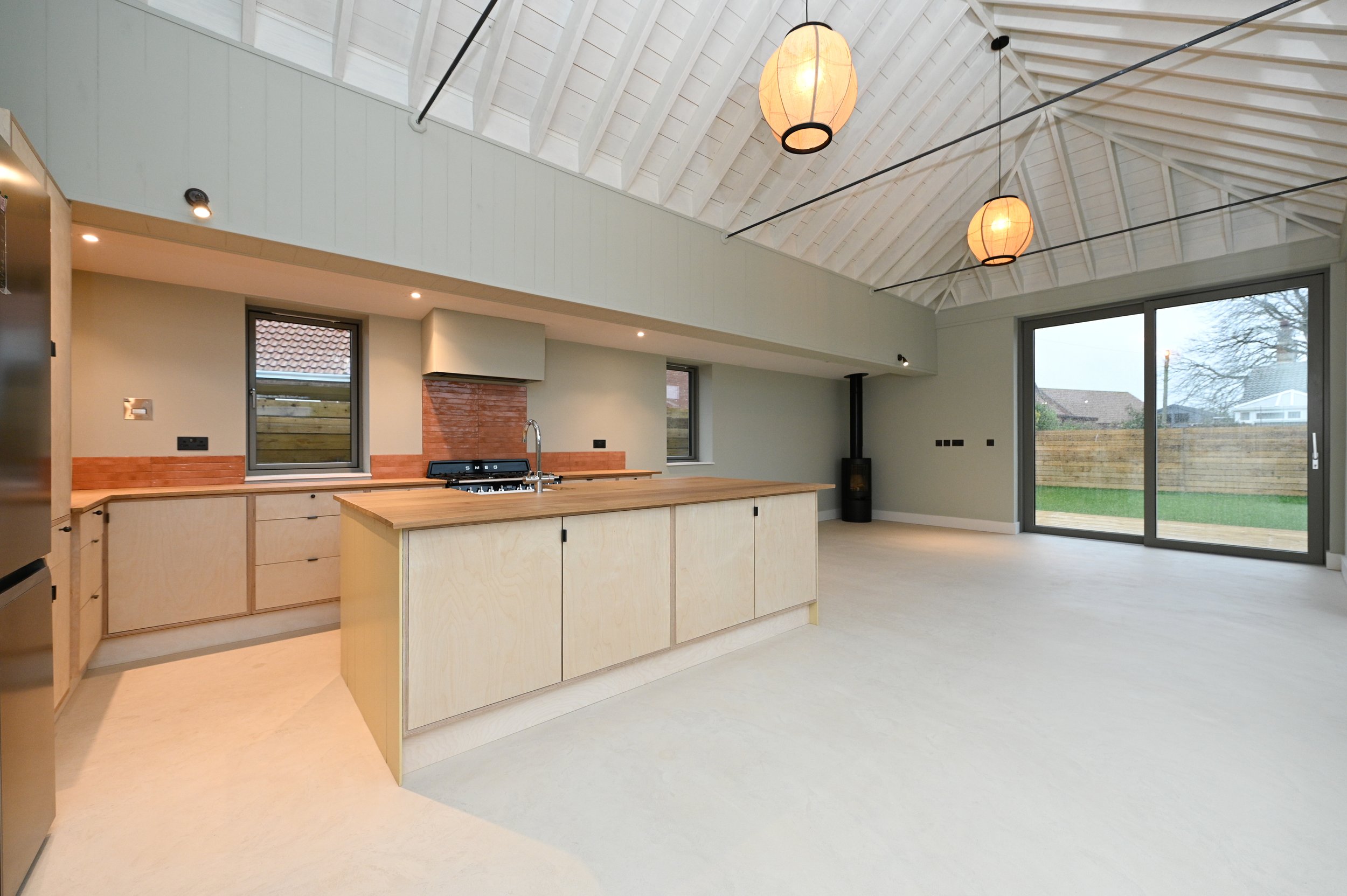 Bright open kitchen with light wood cabinets, a central island, and large sliding glass door leading to a backyard. The ceiling is vaulted with white wooden beams and hanging paper lantern lights.