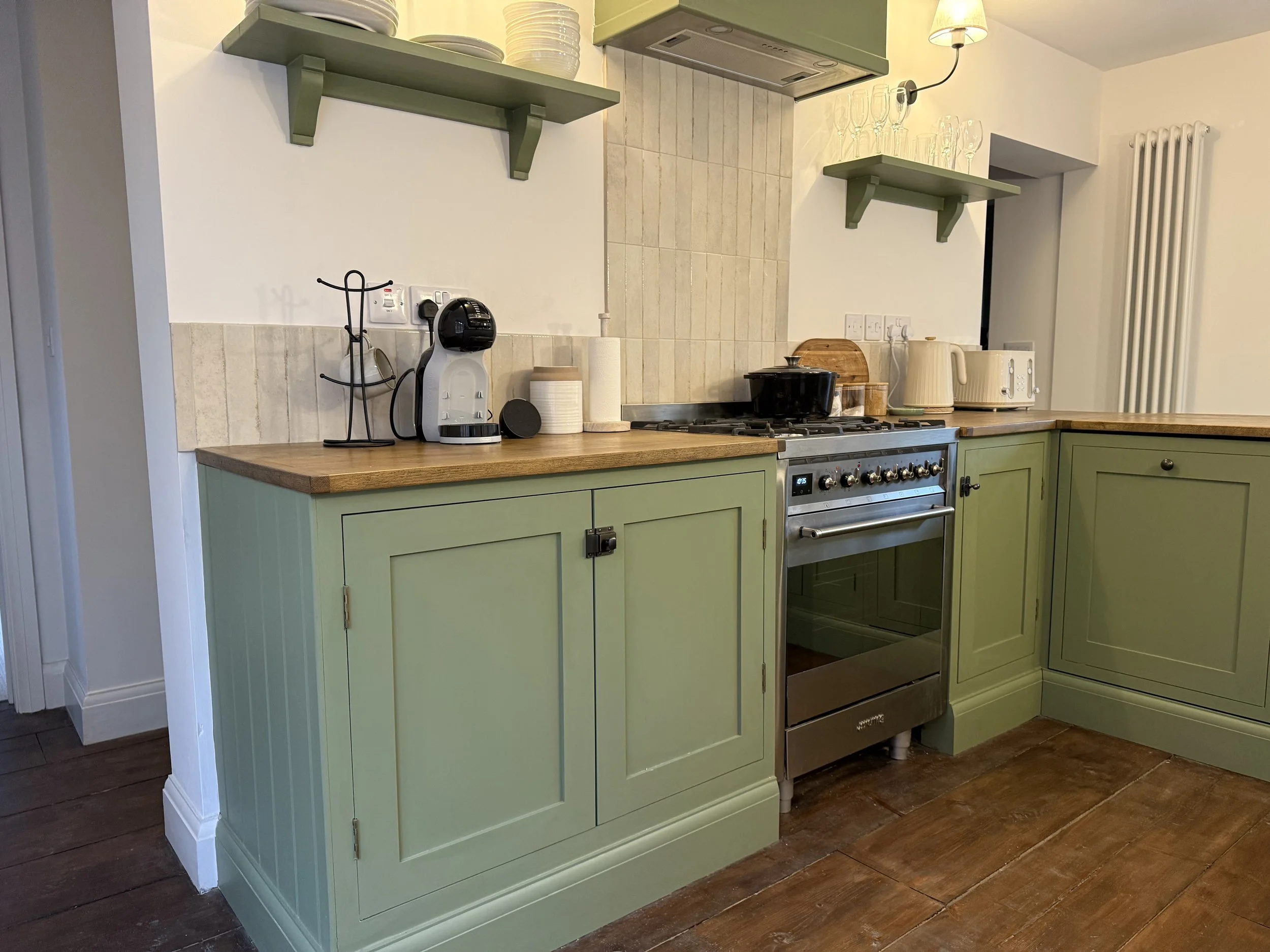 Kitchen with green cabinetry, a wooden countertop, a stove, and various appliances on the counter, including a coffee maker, toaster, and kettle, with green wall-mounted shelves holding dishes and glasses.