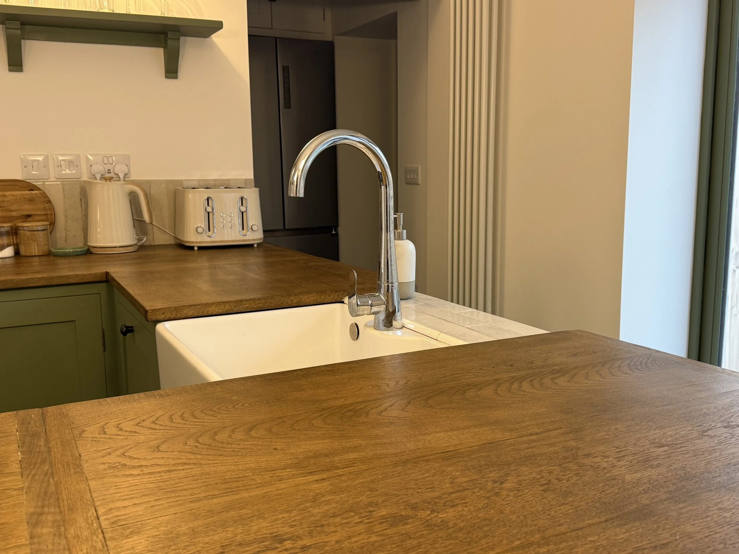 Kitchen with wooden countertops, an off-white sink, a modern faucet, a beige kettle, a toaster, and a fridge in the background. There is a green shelf and a window with a sliding door.