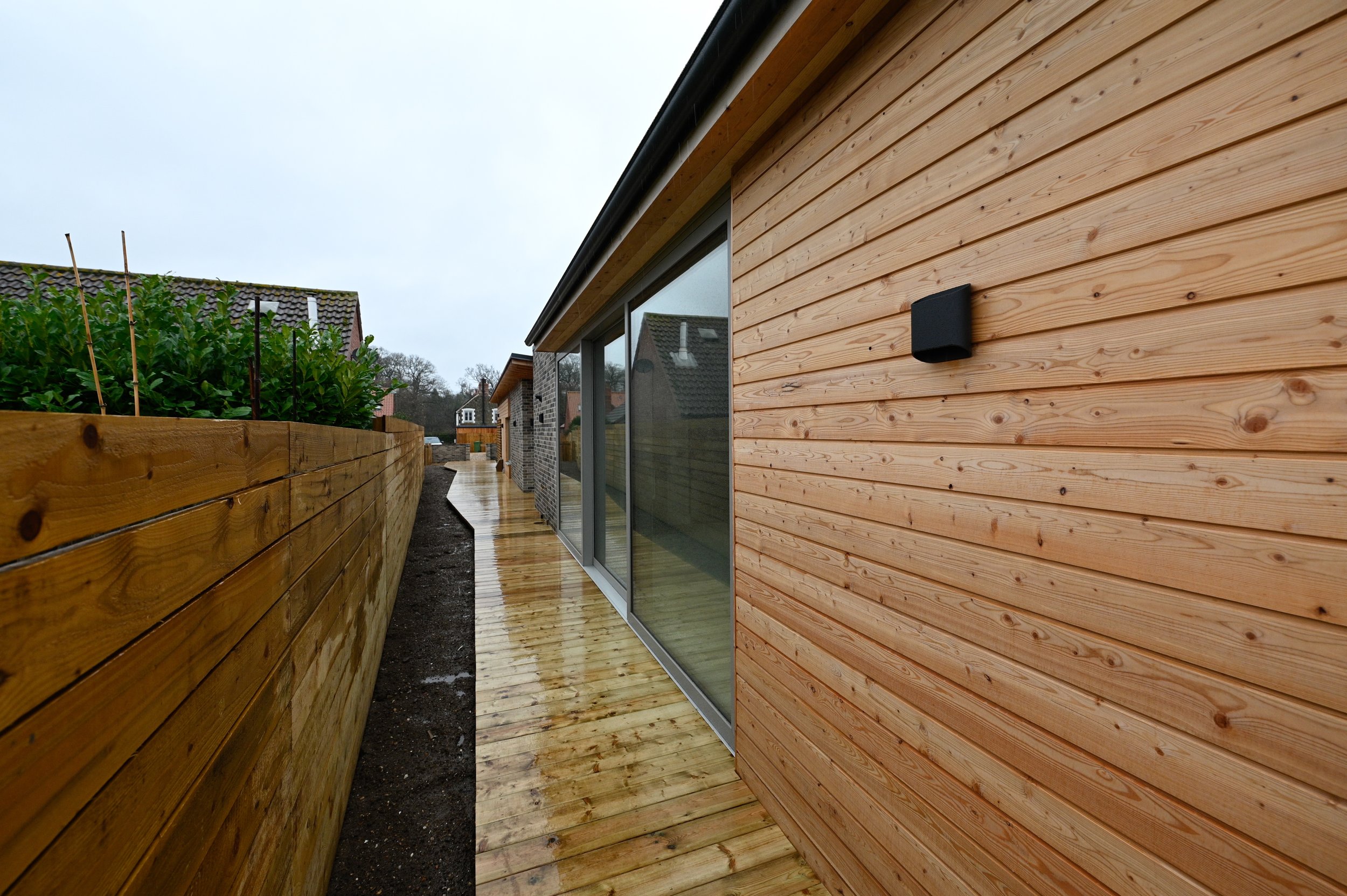 A wooden-sided modern house with large glass sliding doors and a wet wooden deck, bordered by a wooden fence with greenery. Overcast sky.
