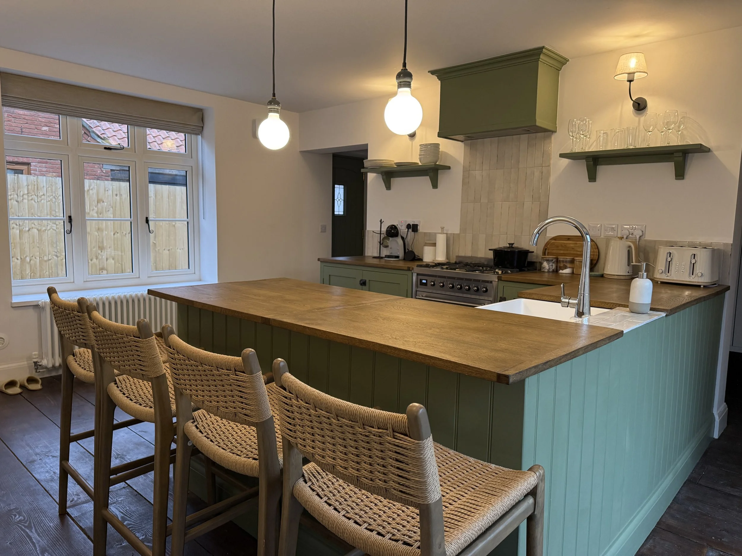 Kitchen with green island, wooden countertop, white backsplash, and open shelving with glassware and dishes. Four wicker chairs are positioned along the island, and two hanging pendant lights illuminate the space.