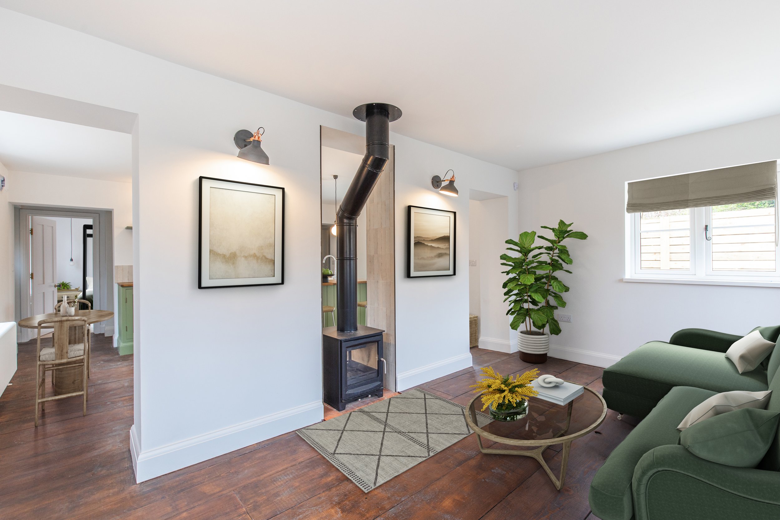 Living room with green sofa, wooden coffee table with yellow flowers, potted plant near window, framed art, wood stove with chimney, hardwood floors, and white walls.