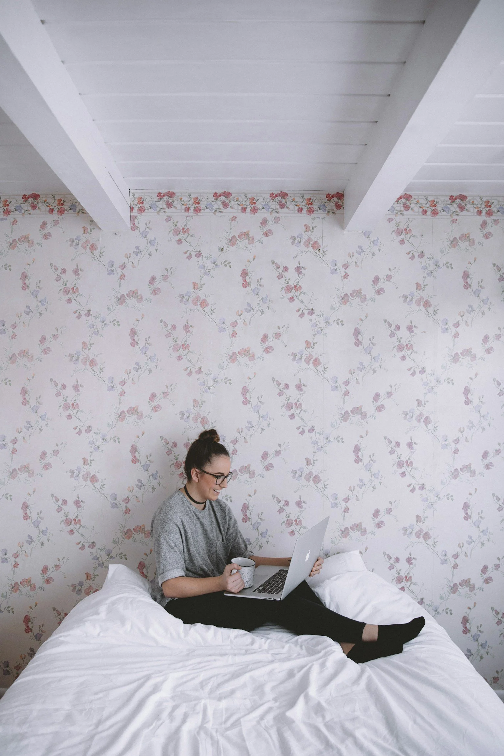 A woman sitting on a bed, working on a laptop while holding a coffee mug, in a room with floral wallpaper and exposed wooden beams on the ceiling.