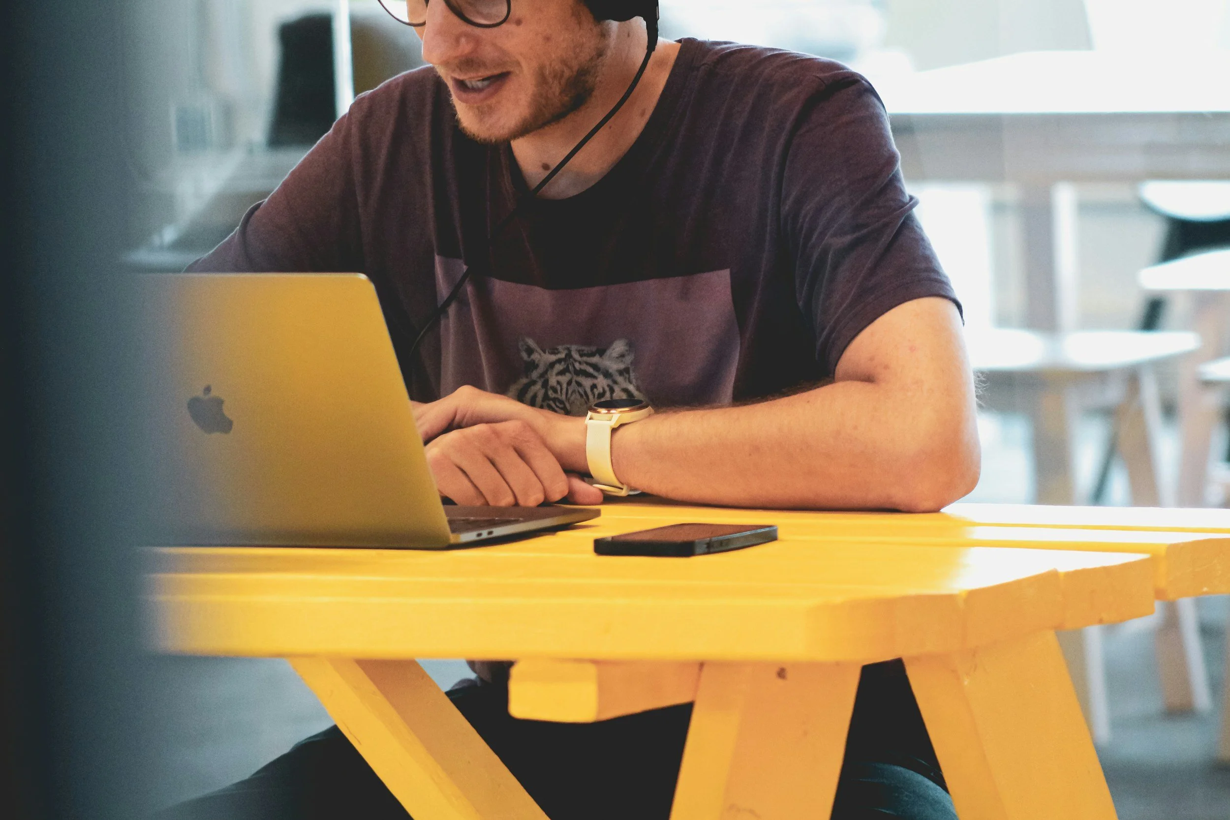 A young man wearing glasses, a graphic t-shirt with a tiger, and a yellow watch is sitting at a yellow table, working on a laptop.