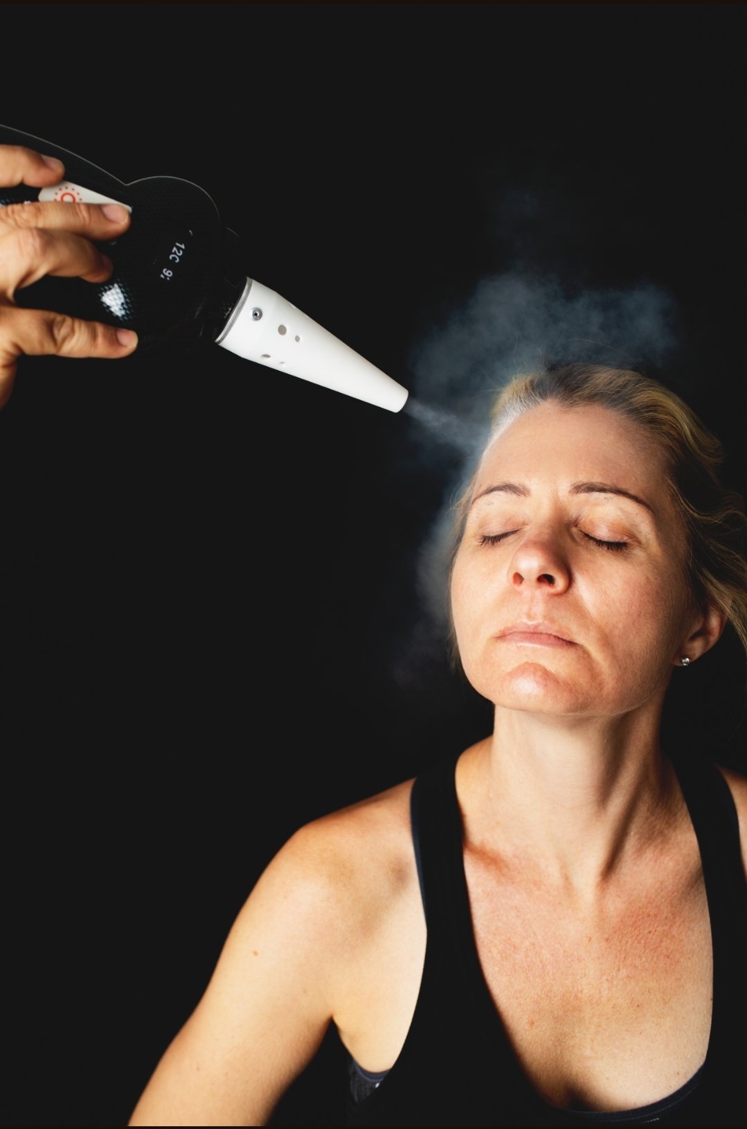Woman with eyes closed as a handheld vaporizer is used on her forehead, emitting vapor over her face against a black background.
