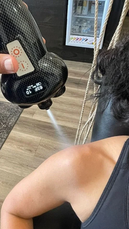 A person with black hair getting a shoulder massage while a hand sprays water from a black misting device in a room with wooden floors and a vending machine in the background.