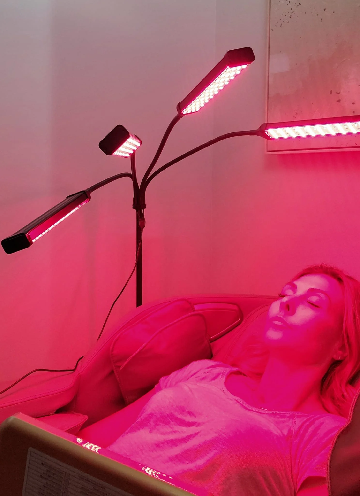 A woman lying on a bed under red LED light therapy devices, with her eyes closed and wearing a pink top.