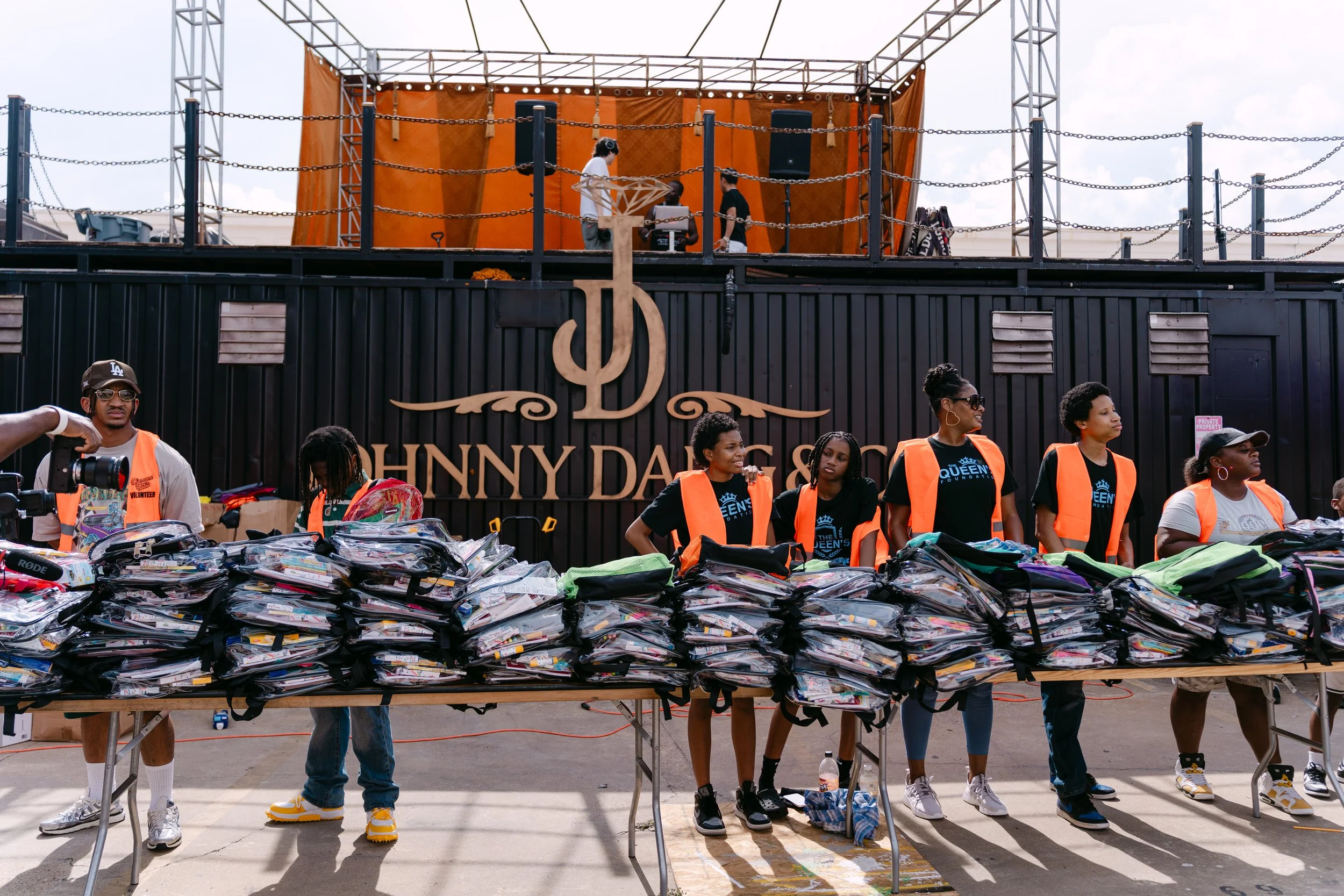 People standing behind tables filled with bags, in front of a black wall with a logo and the words "Johnny Dangers". Some individuals are wearing orange safety vests and black T-shirts with a blue crown emblem.