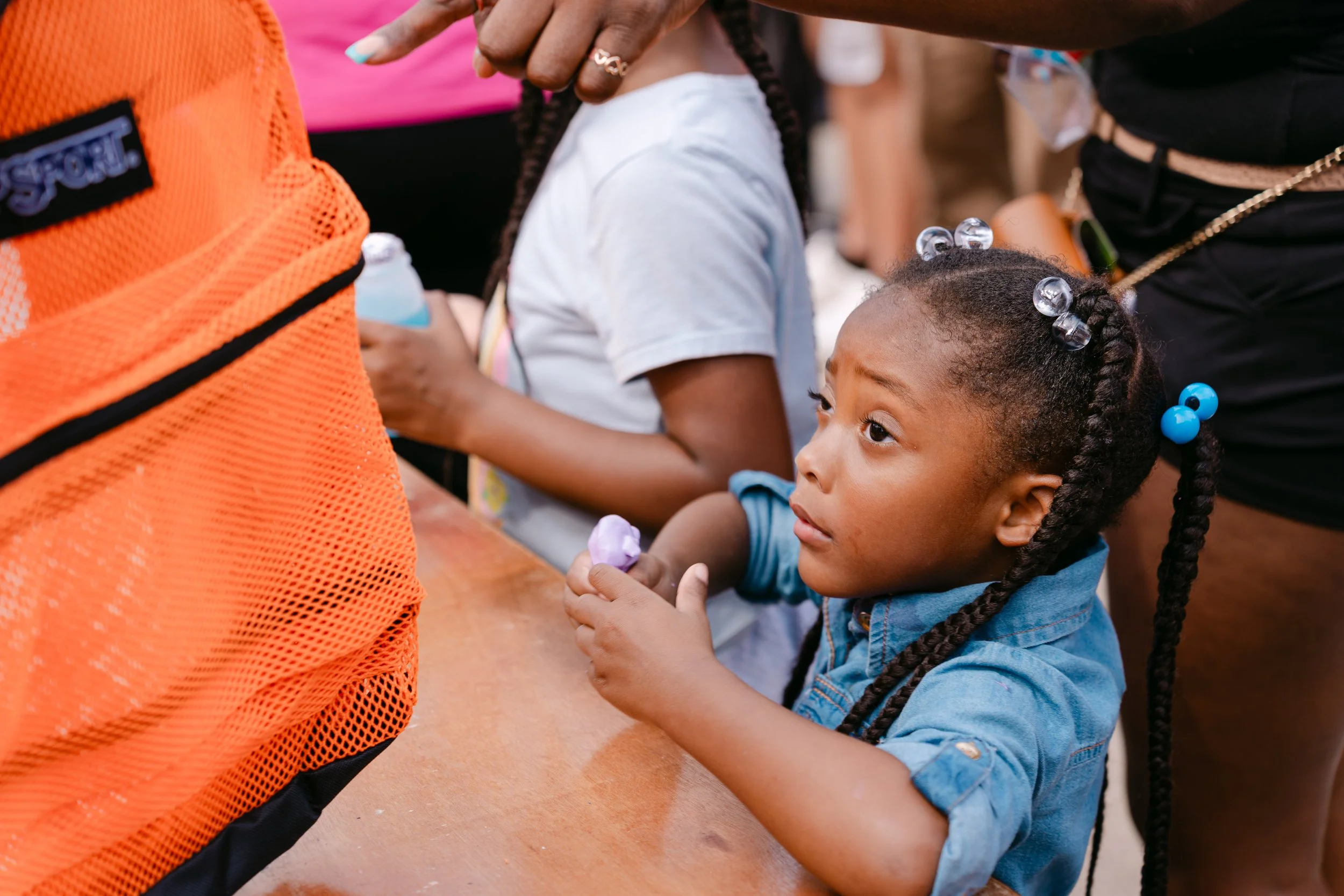 A young girl with braided hair and blue beads, sitting at a table and holding a small object, surrounded by other children and adults.