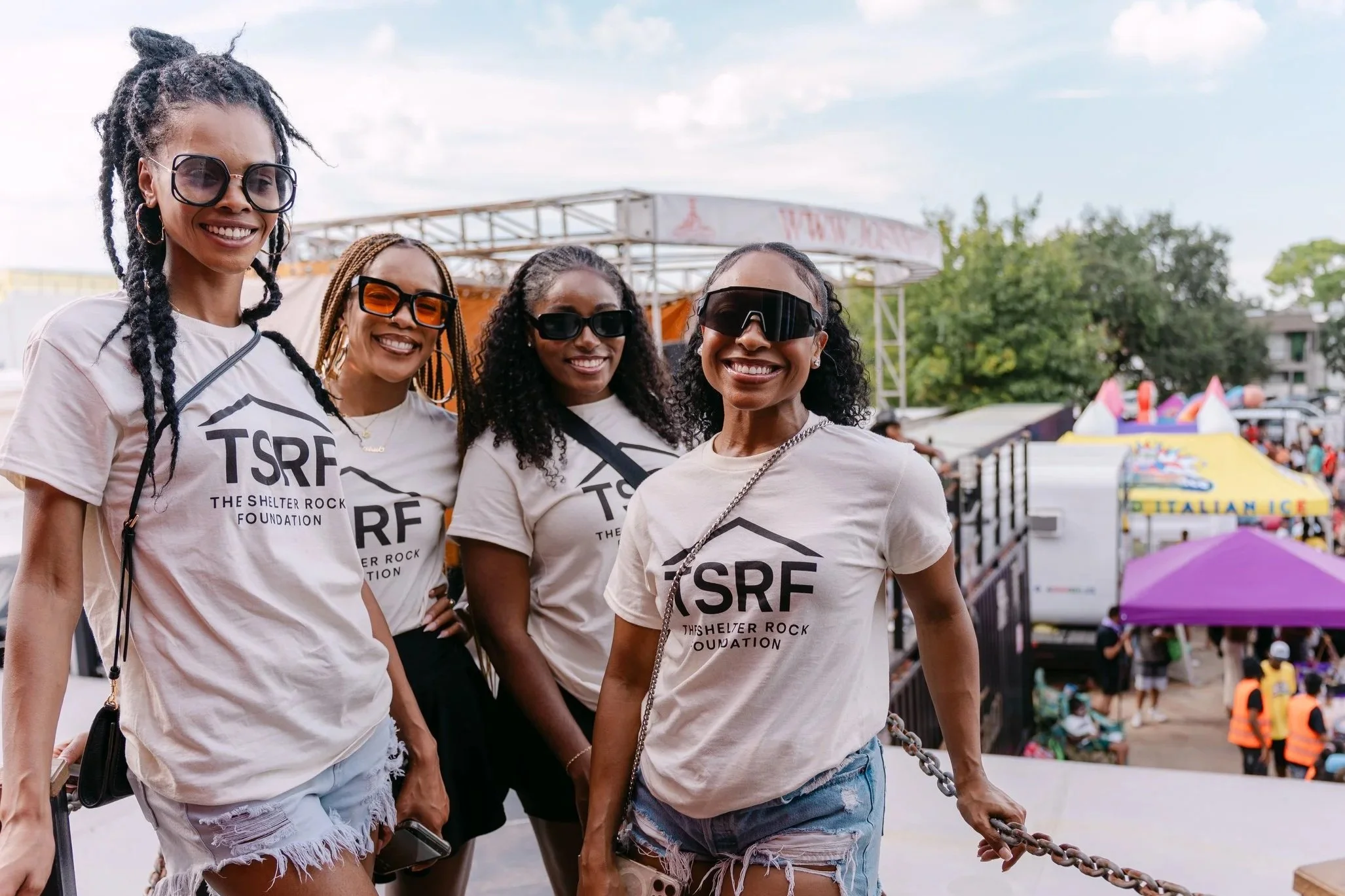 Four women smiling at an outdoor event, wearing matching T-shirts with 'TSRF The Shelter Rock Foundation' printed on them, all wearing sunglasses, with tents and crowds in the background.