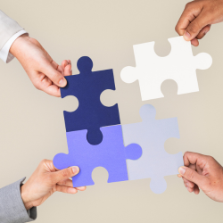 Four hands each holding a puzzle piece, connecting to form a square, with colors navy blue, light blue, white, and light gray.