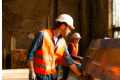 Construction worker wearing a safety vest and helmet working with blueprints inside a building under construction.