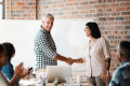 A man and woman shaking hands in a casual office setting with colleagues at a table