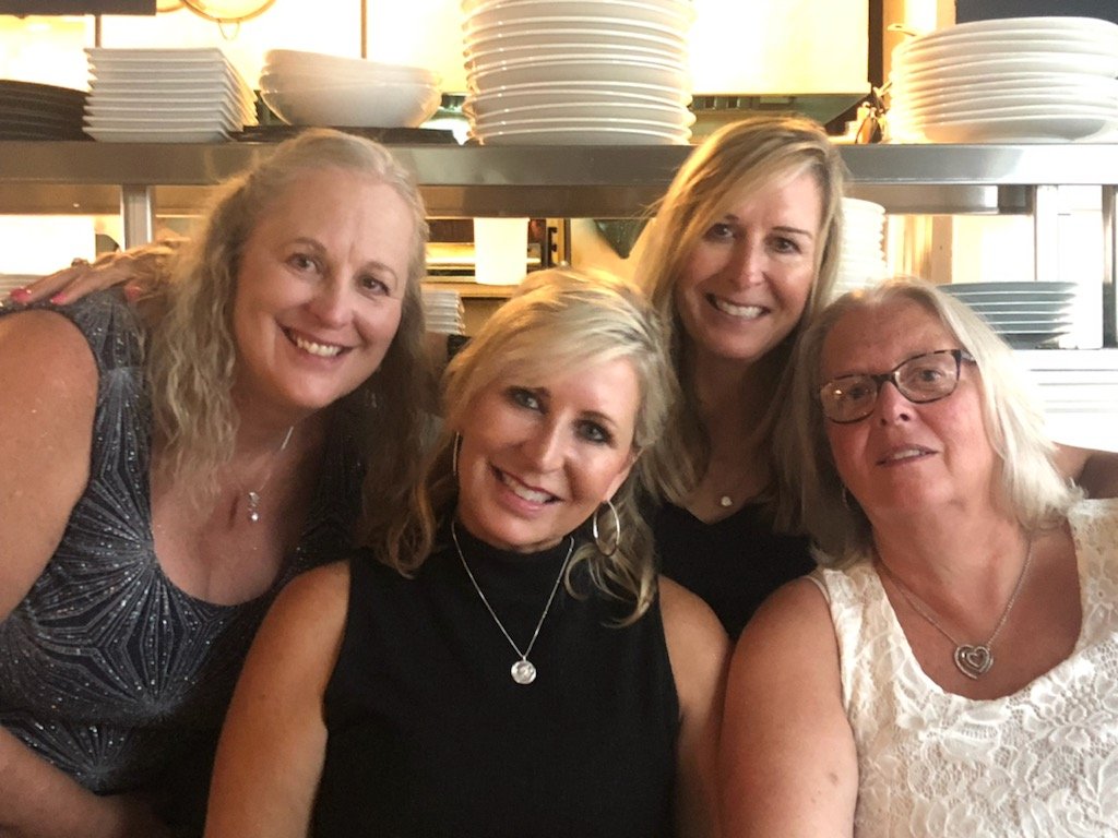 Four women posing together in a restaurant or kitchen with stacked white plates and bowls in the background.