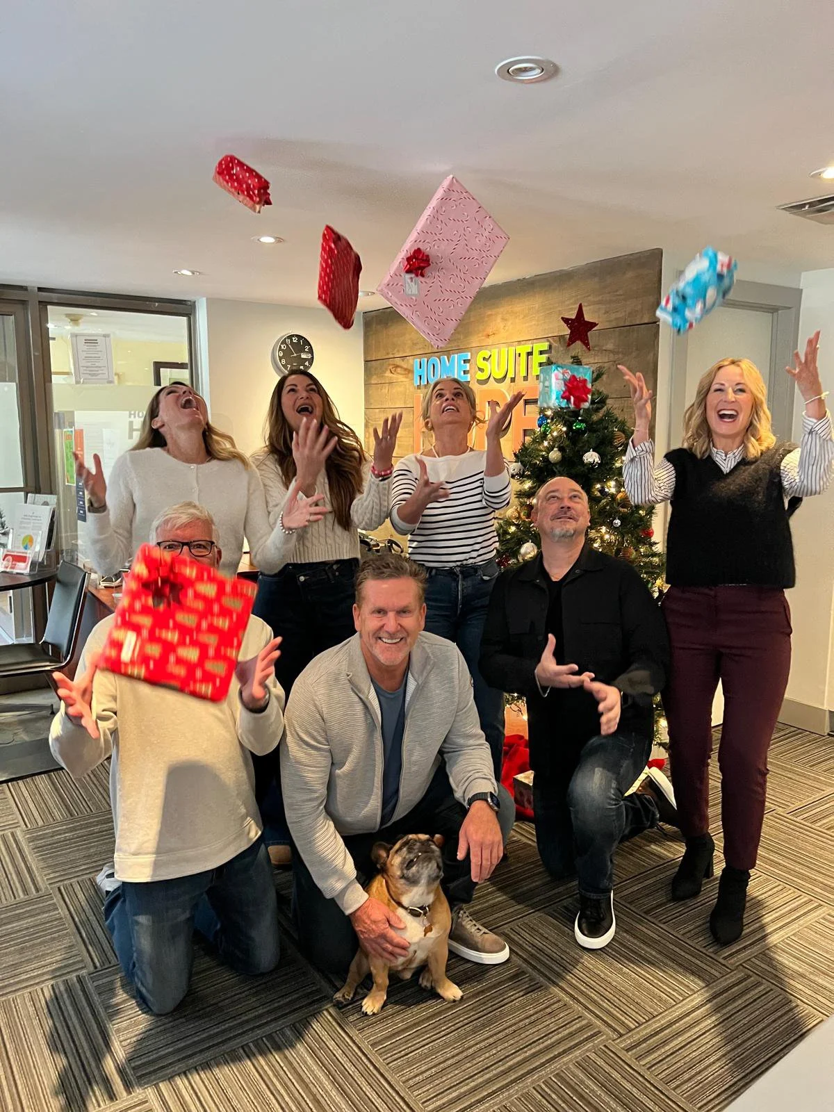 The Dan Cooper Group had just finished wrapping gifts they purchased for families at Home Suite Hope and celebrated by taking some photos and were throwing the wrapped gifts in the air, with a decorated Christmas tree in the background.
