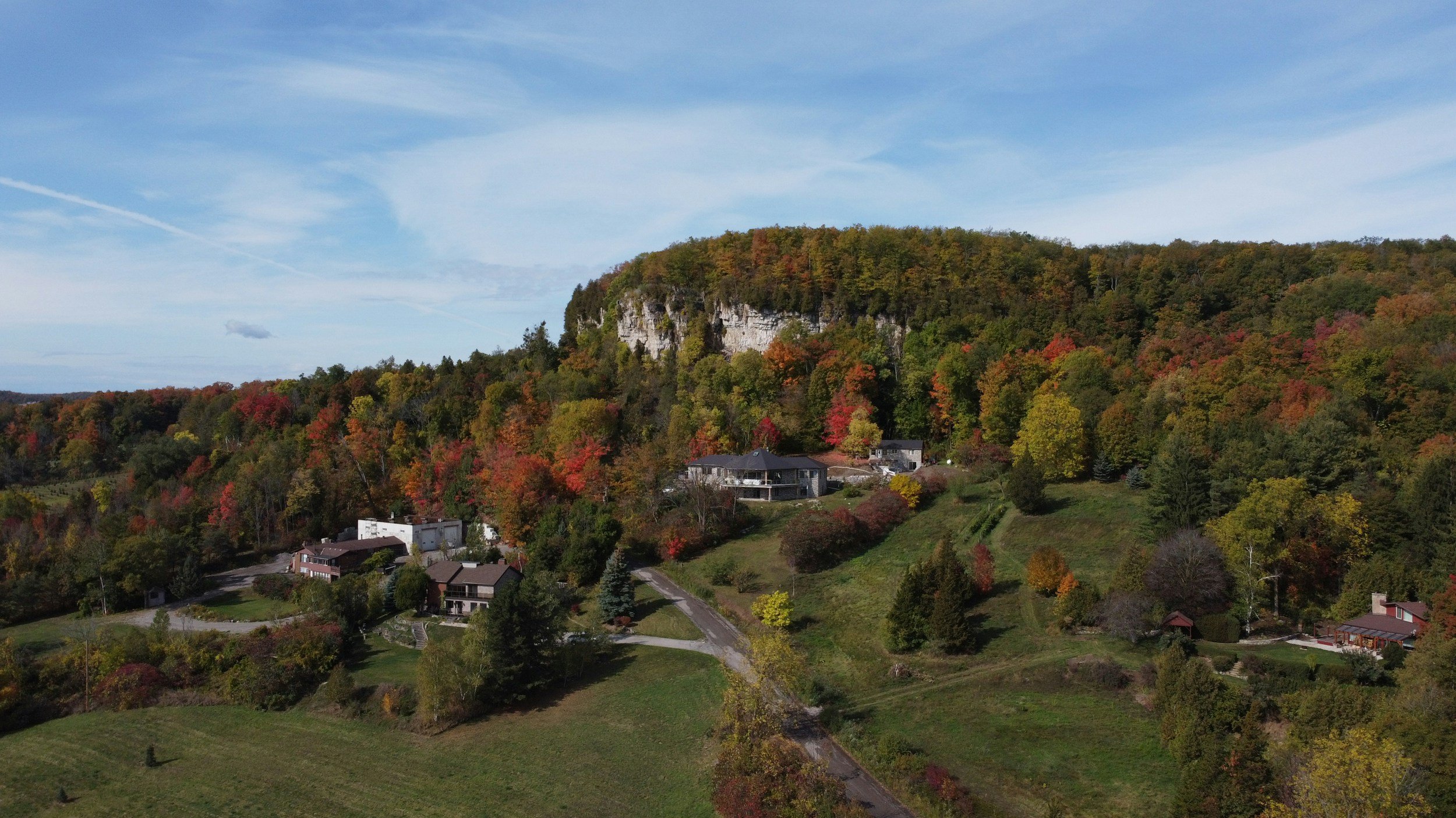 A panoramic view of a hillside covered with autumn foliage, with houses at the base, a rock cliff on top, and a blue sky with some clouds.