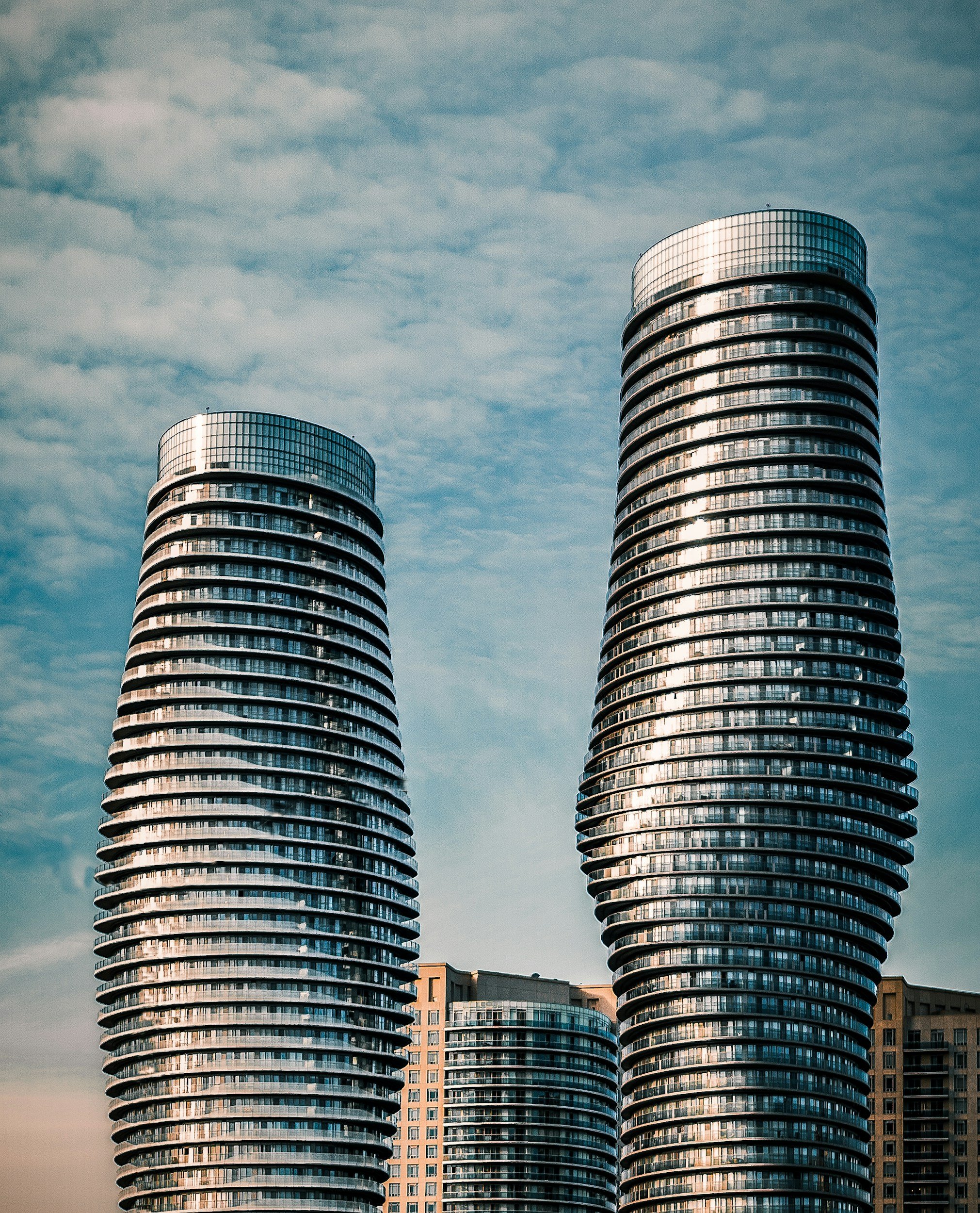 Two modern, curved high-rise buildings with glass exteriors, against a cloudy sky, with lower structures at the base.