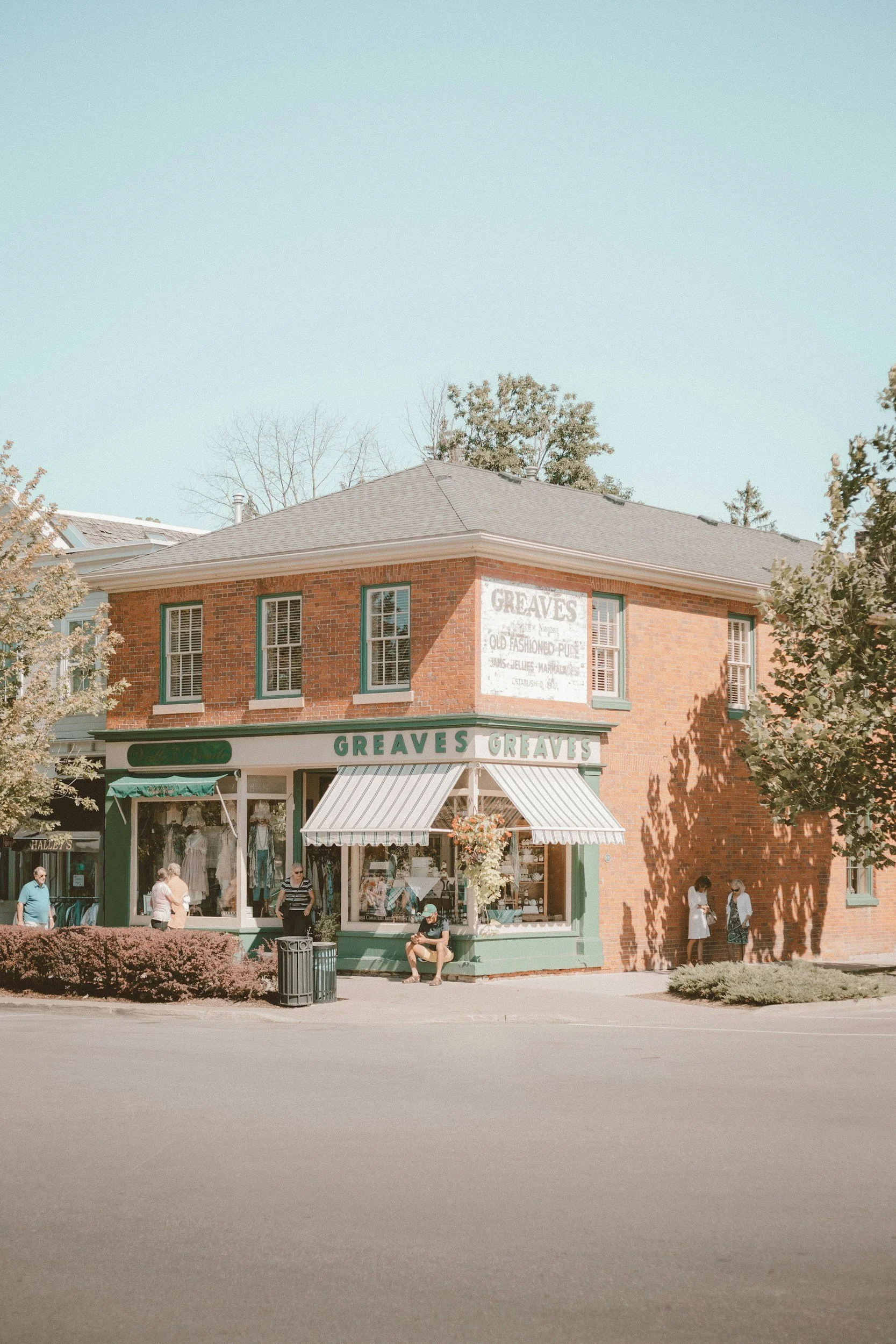 A two-story brick building with a green storefront named "Grevés" on a city street. The shop has large display windows, a green awning, and an upper sign with faded text. People are walking and sitting outside, with trees and a clear sky in the background.