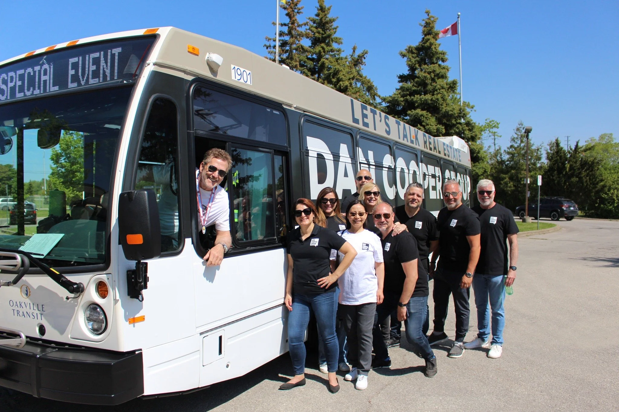 Group of people standing next to a white transit bus with 'Let's Talk Real Estate' written on top and 'Dan Cooper Group' on the side, outdoors with trees and a flagpole with the Canadian flag in the background.