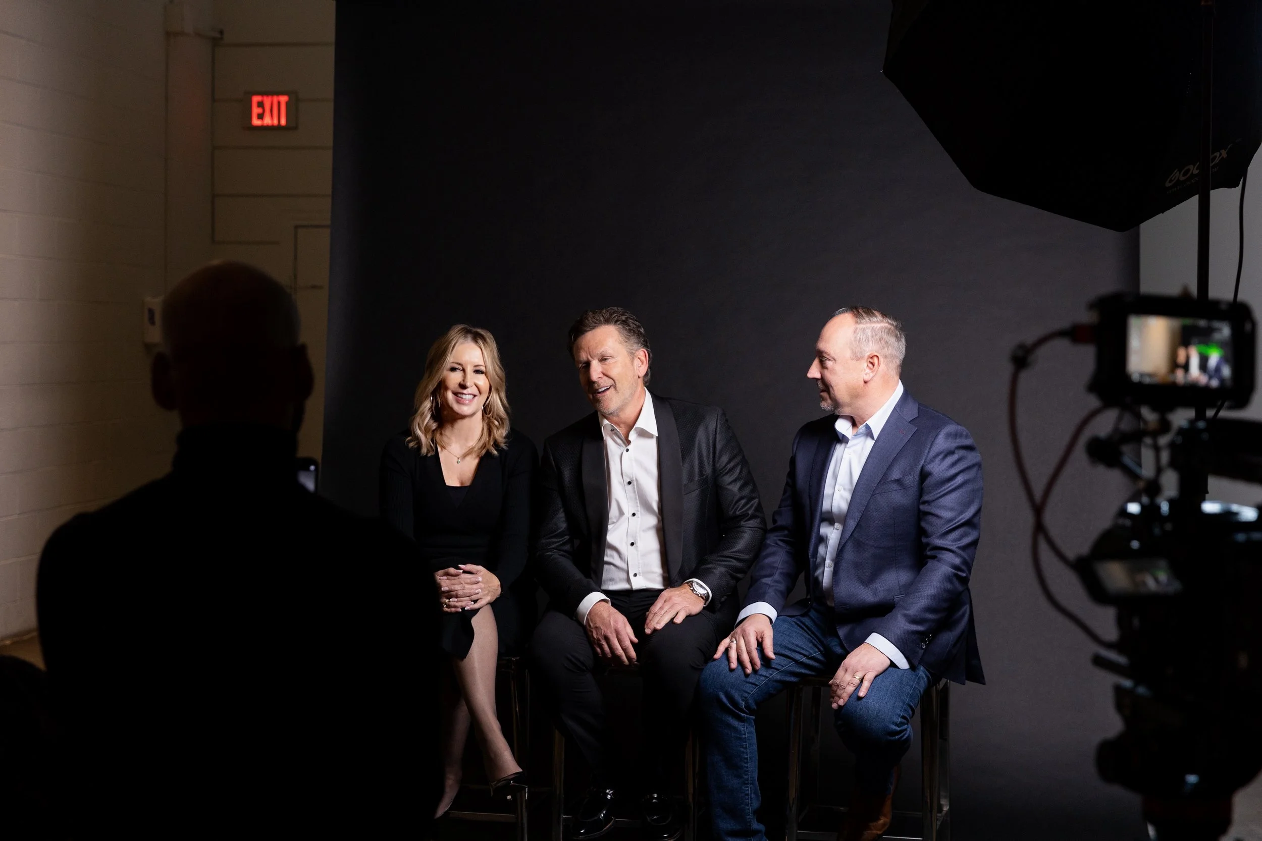 Four people, two men and two women, are sitting and smiling in a black background during a photo shoot. A camera and lighting equipment are visible in the foreground.