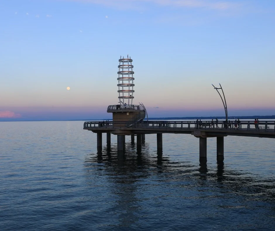 A pier extending into a calm body of water with modern curved and spiral structures, a tower with a spiral design, and people walking on the pier. The sky is clear with a visible moon.