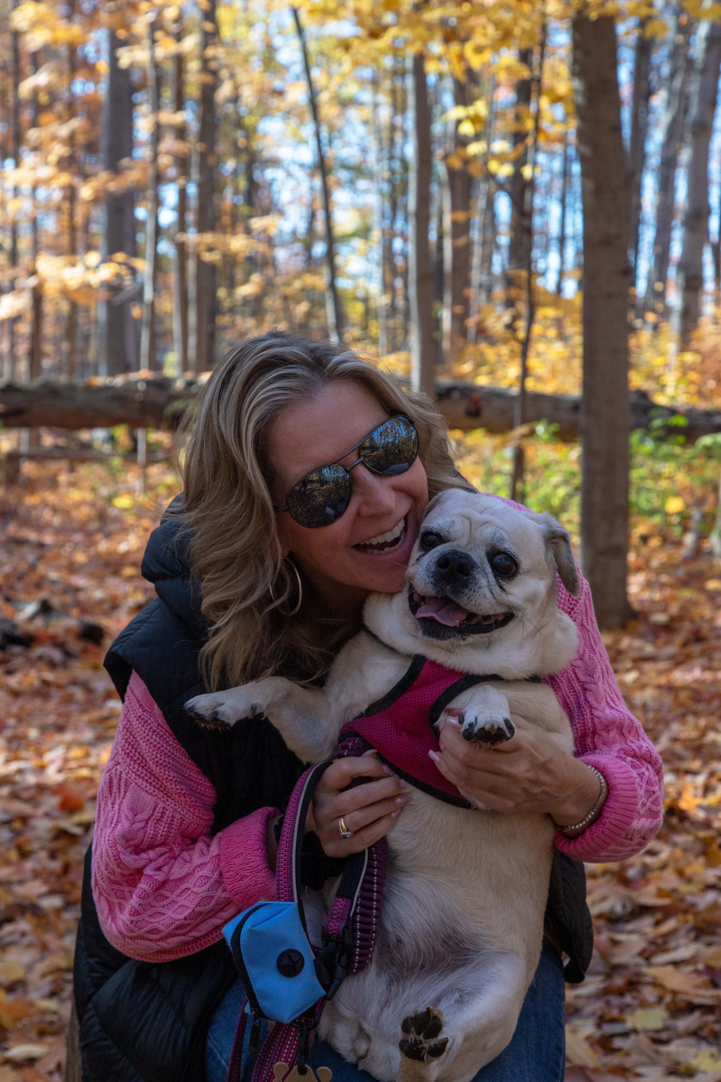 A woman wearing sunglasses and hiking gear holding a happy, light-colored dog in a fall forest with yellow and orange leaves.
