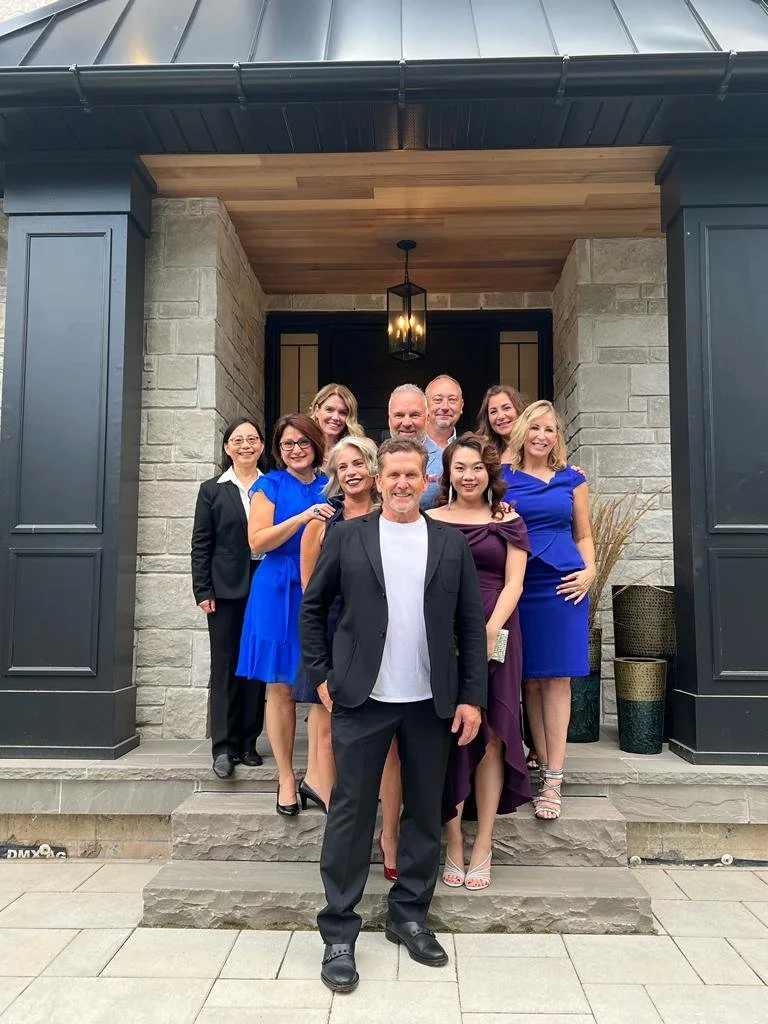 A group of ten well-dressed people, including eight women and two men, posing for a photo on the steps of a house with gray siding and stone walls, during daytime.