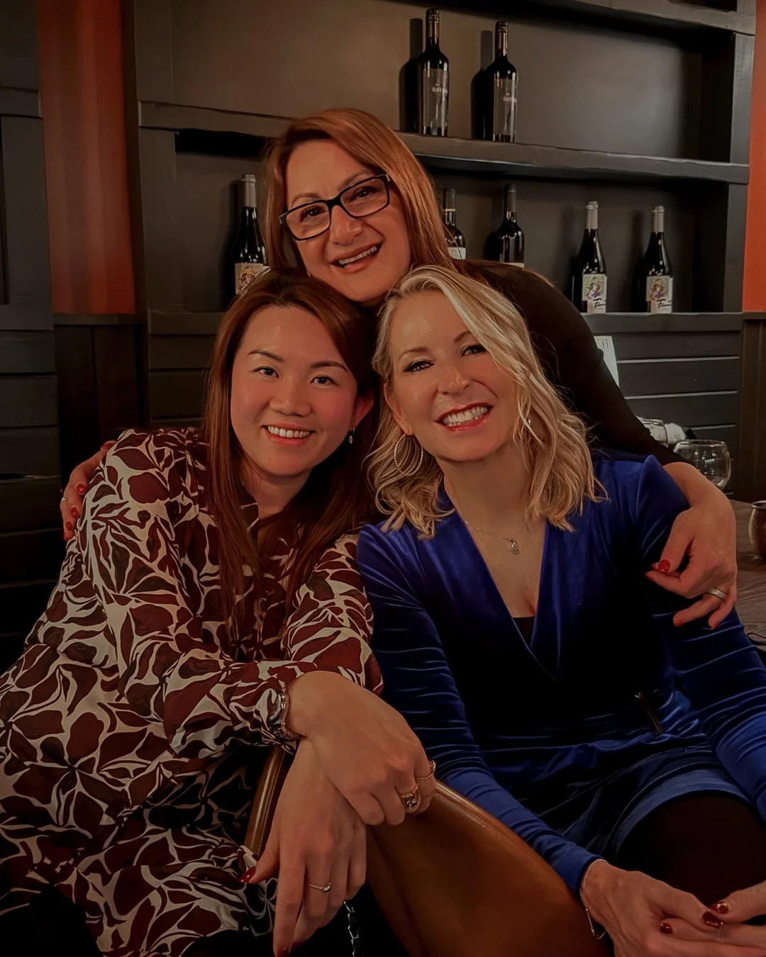Three women posing together indoors, smiling, with a dark wooden shelf holding wine bottles in the background.