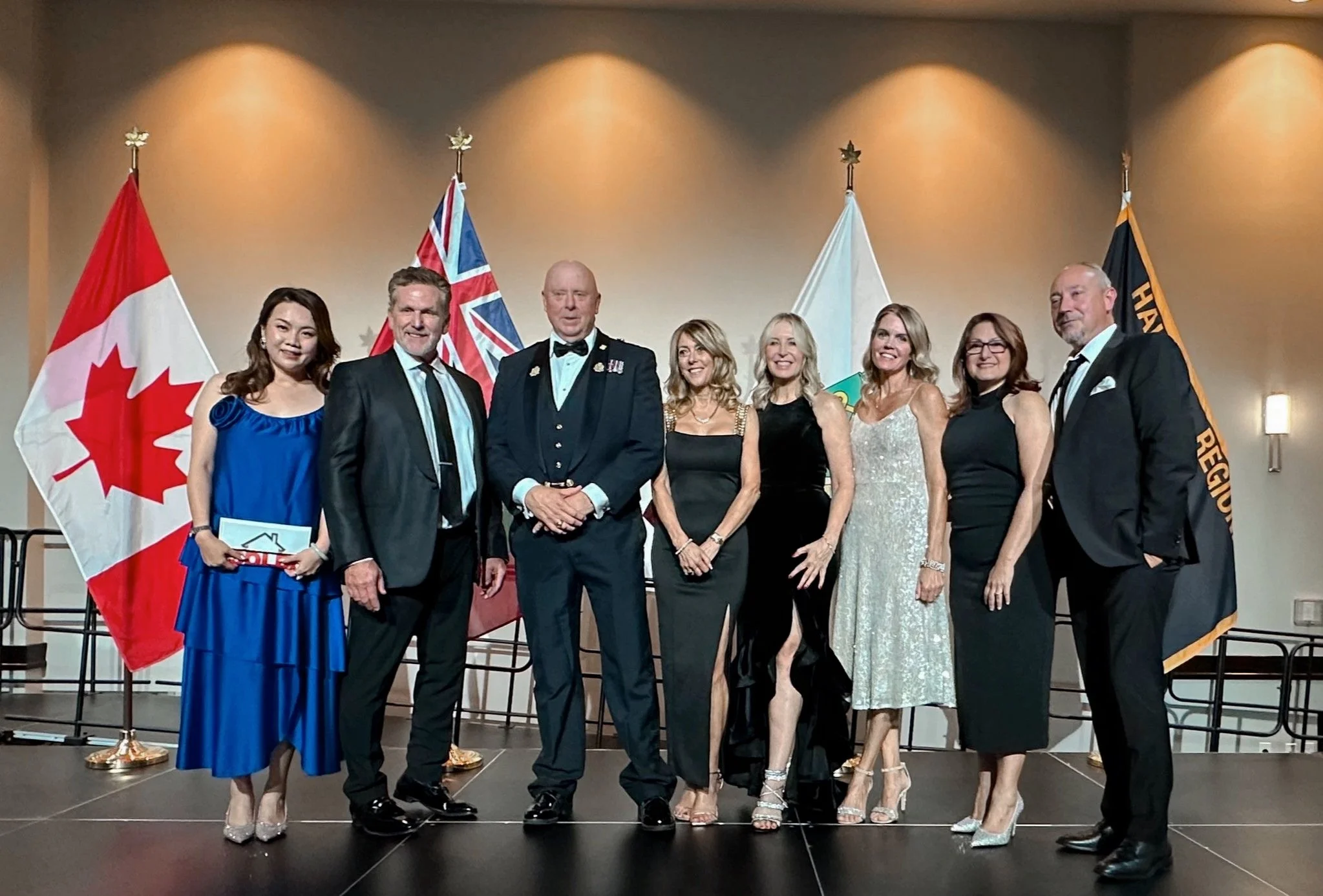 Group of nine formally dressed people standing on stage with flags in the background, including the Canadian flag, a Union Jack, and a white flag. The group is smiling, and the setting appears to be an award or recognition ceremony.