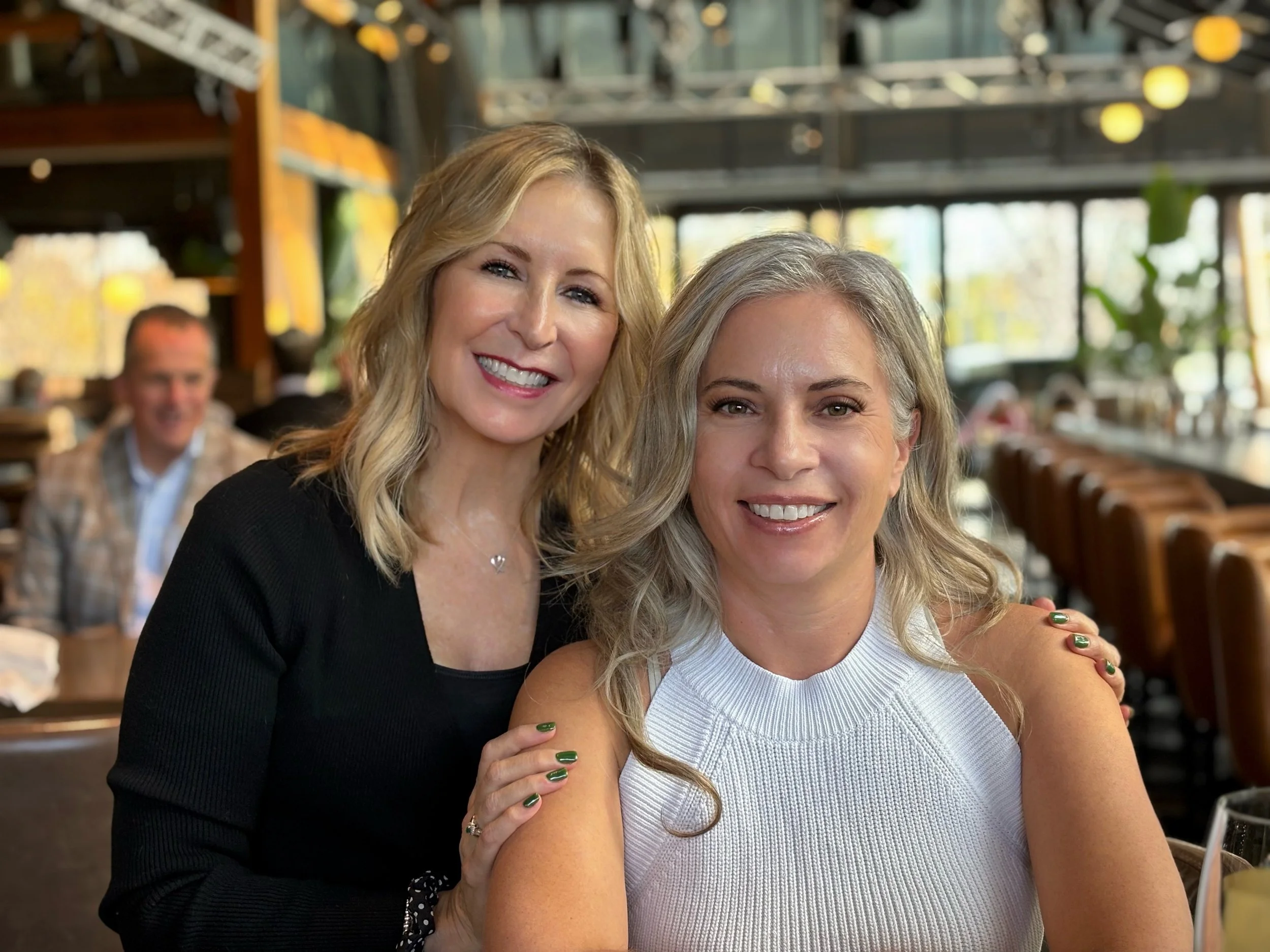 Two women smiling and sitting close together at a restaurant or cafe. One woman has blonde hair and is wearing a black top, the other has gray hair and is wearing a white sleeveless top. There are other people and a modern, well-lit interior in the background.