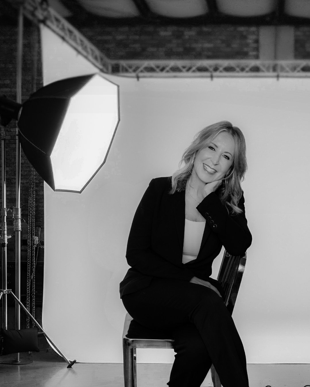 Kathy Wood sitting on a chair in a photography studio, smiling, with professional lighting equipment and a white backdrop.