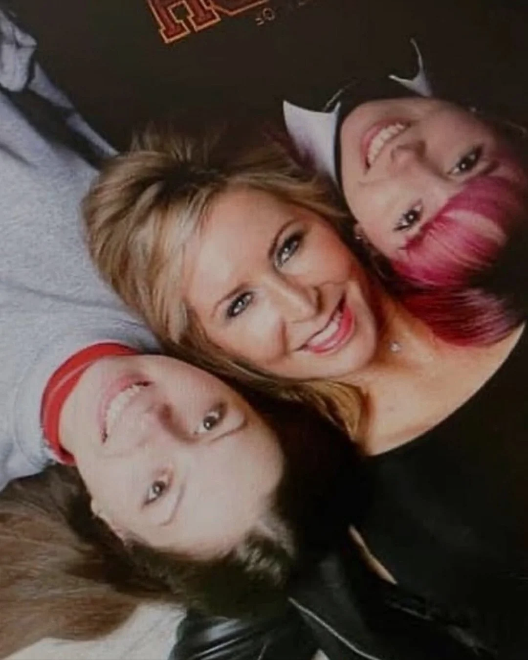 Three women lying down together, smiling at the camera. They have different hair colors: blonde, pink, and brown.
