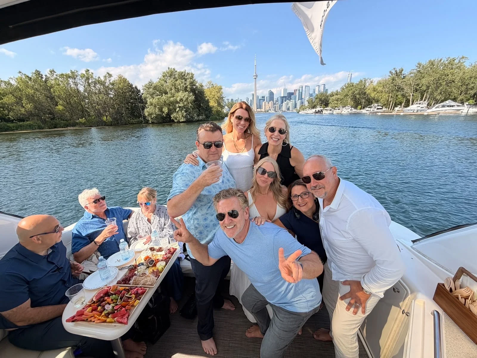 A group of nine adults enjoying a boat party on a sunny day with a city skyline and river in the background, featuring the CN Tower. They are smiling, wearing sunglasses, and have a table with snacks and drinks on the boat.