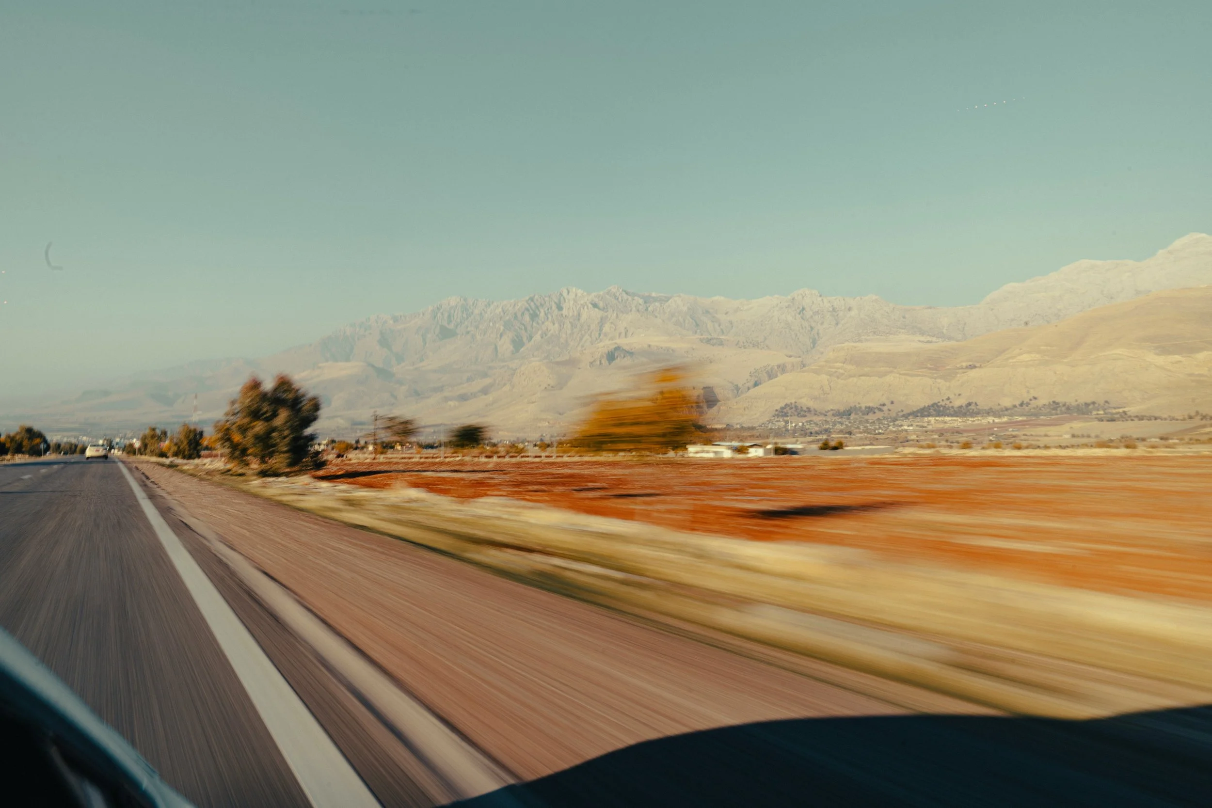 Blick auf eine asphaltierte Autobahn mit gelben Linien, umgeben von Herbstbäumen und einer Wüstenlandschaft im Hintergrund, unter klarem blauen Himmel.