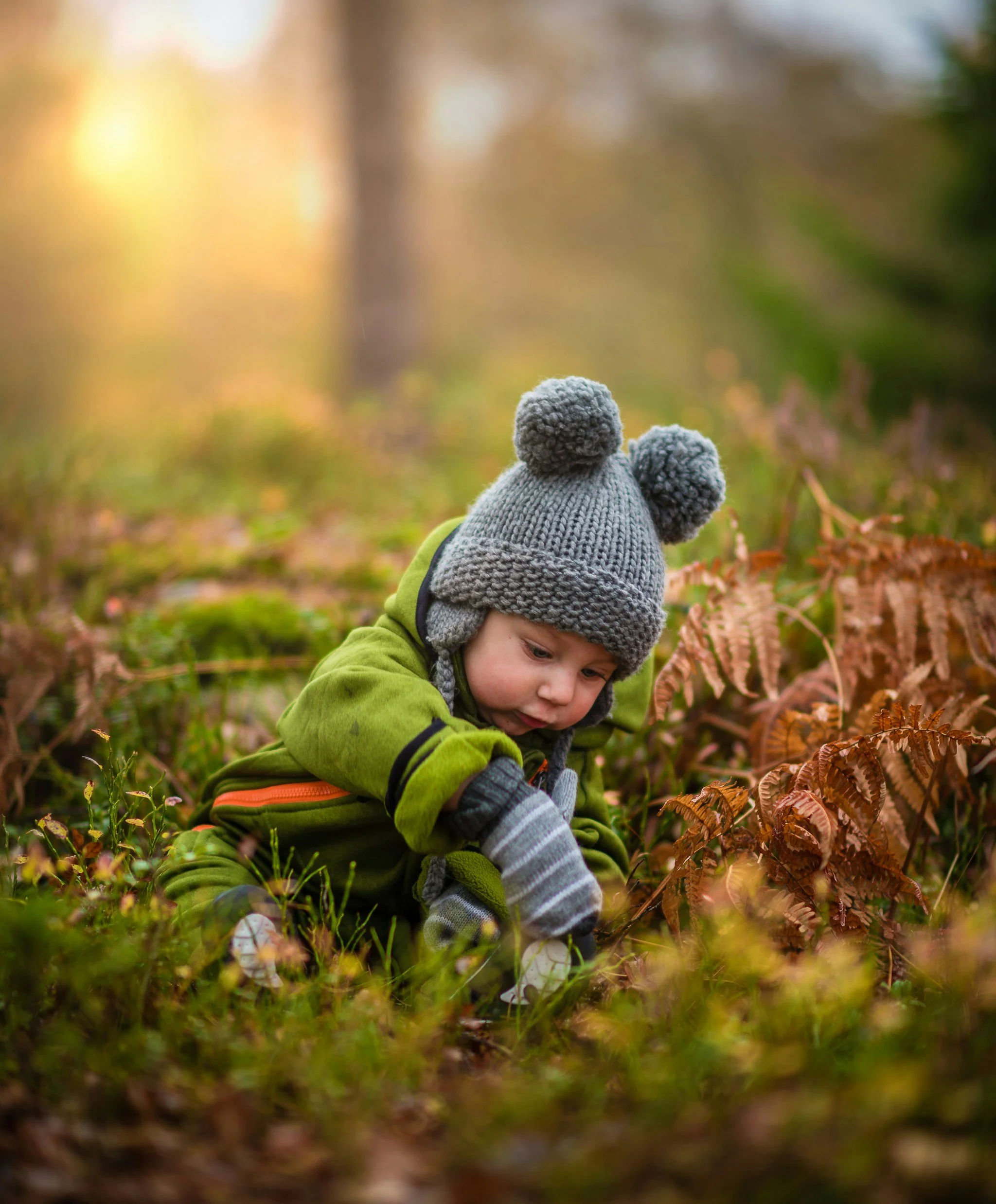 A young child wearing a gray knit hat with pom-poms, a green jacket, and gray gloves, crouching in a forested area with fallen leaves, looking at the ground.