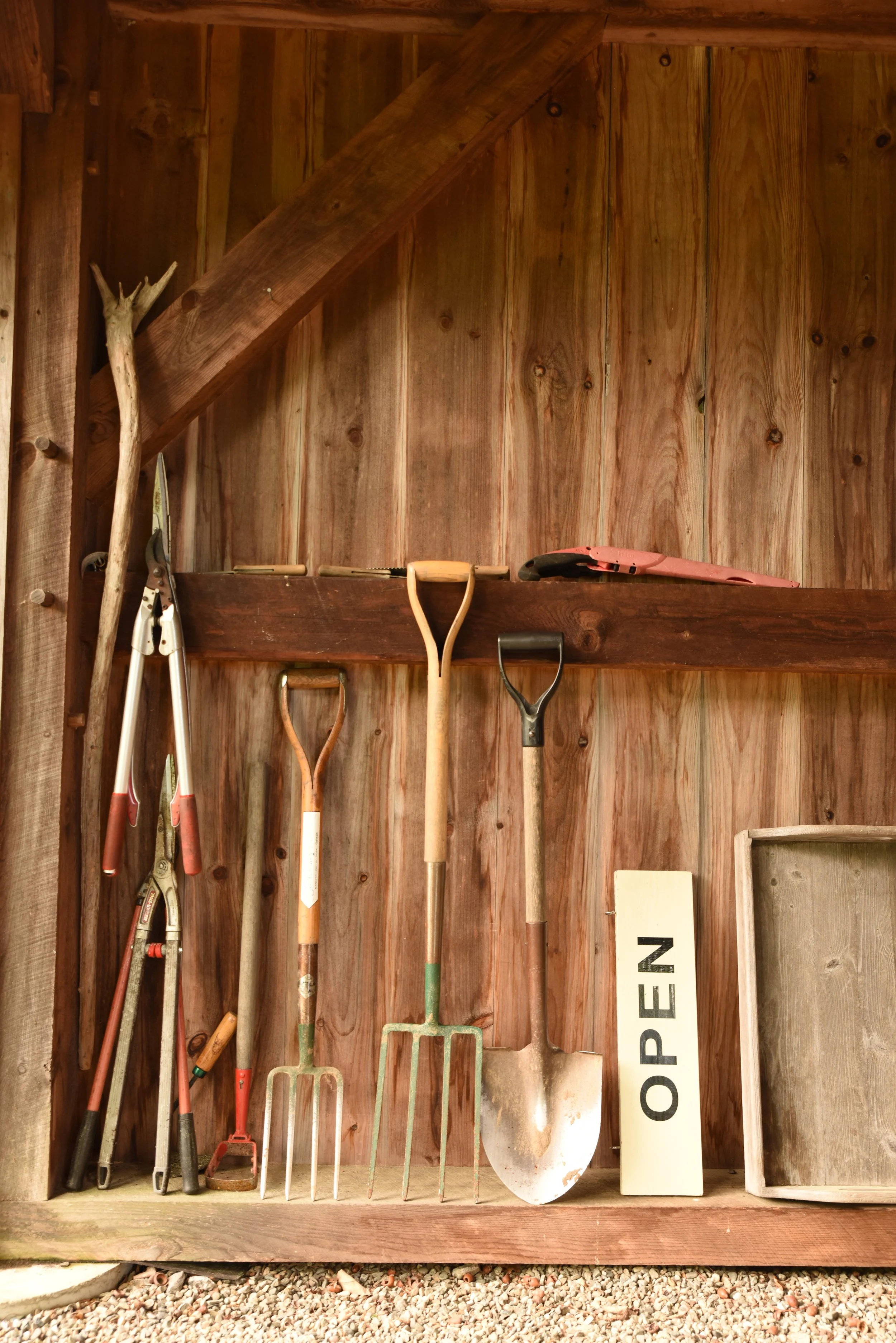 Wooden shed wall with gardening tools hanging, including pruning shears, small rake, large garden fork, shovel, and a wooden frame, with a sign that says 'OPEN'.