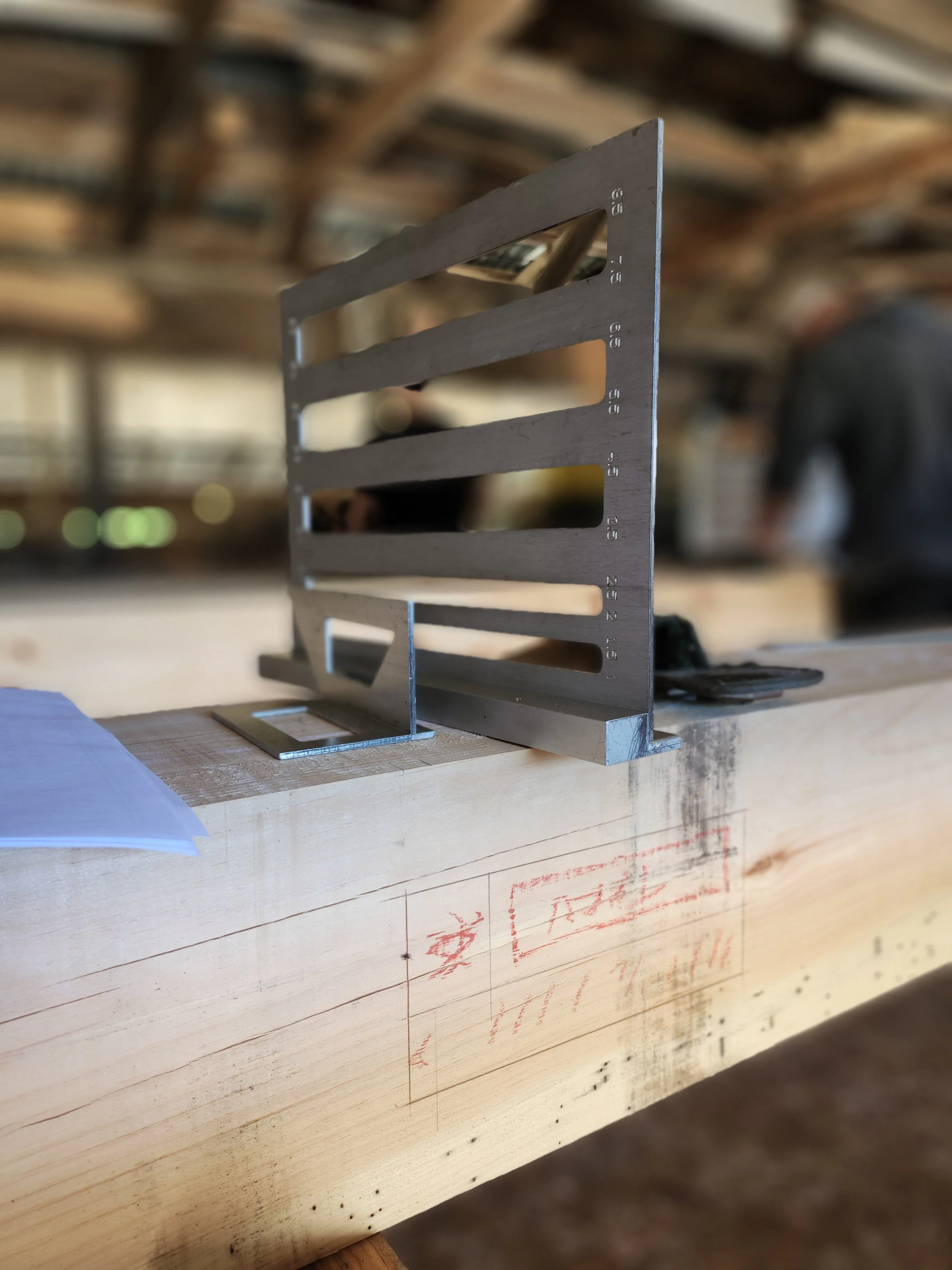 A metal framing square sitting on a wooden workbench in a woodworking shop, with a piece of paper and some drawings on the wood surface and blurred background of workshop setting.
