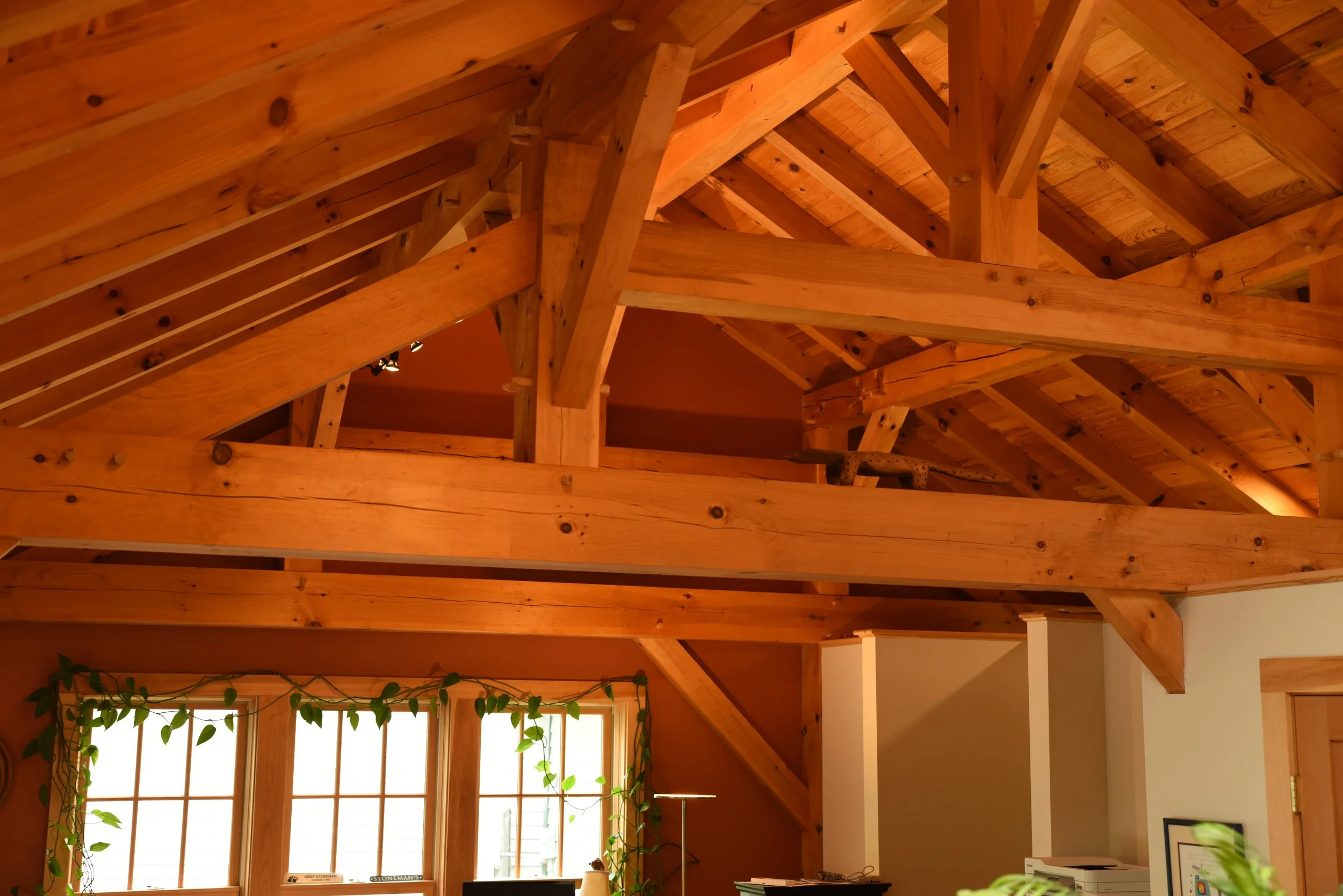 Interior view of a wooden attic with exposed beams, a window with plants, and a white wall with a framed picture.